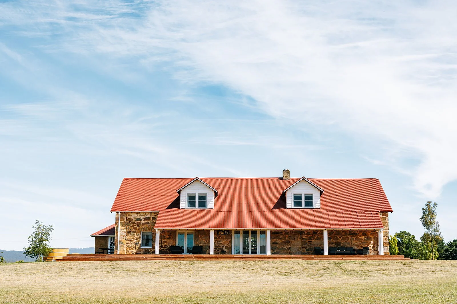 A large house with stone walls, a red metal roof, white-framed windows, and a porch with outdoor furniture, set against a grassy field and a partly cloudy sky.