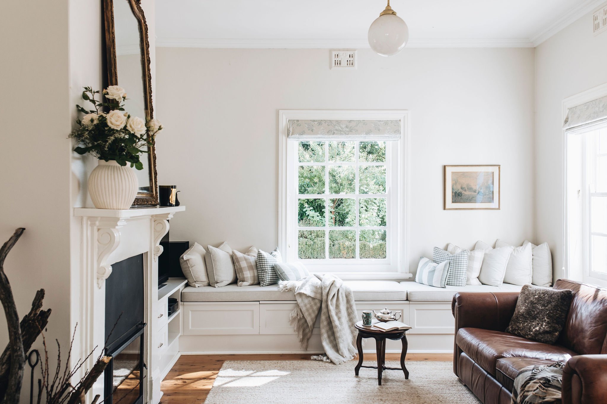 Bright living room with a large window seat, decorative pillows, and throw blanket, brown leather armchair, small wooden side table with teacup and book, and a white fireplace with a floral vase and mirror above.