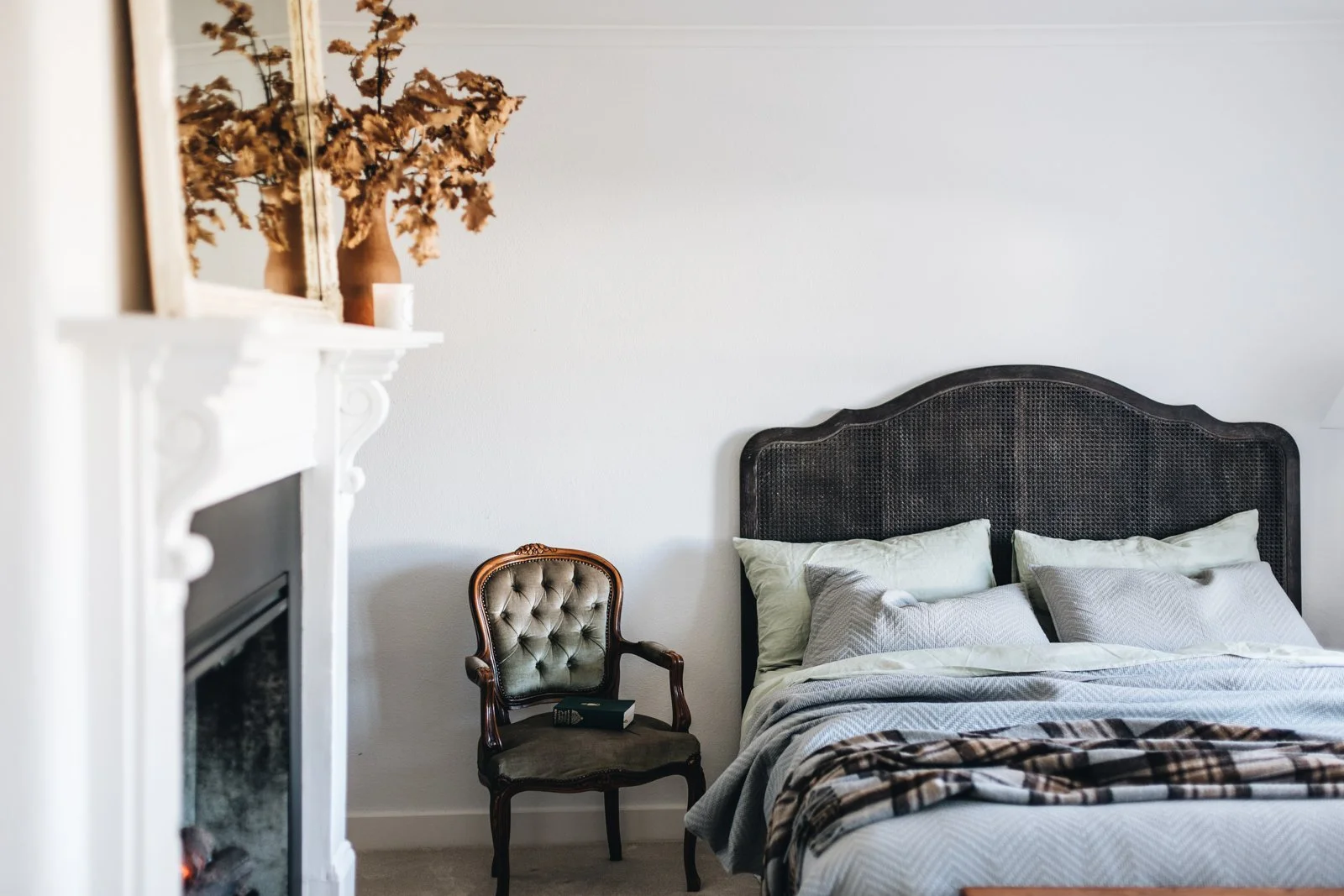 Bedroom with a bed and pillows, a vintage chair with a book on it, a white fireplace mantle with dried flowers and a mirror above it.