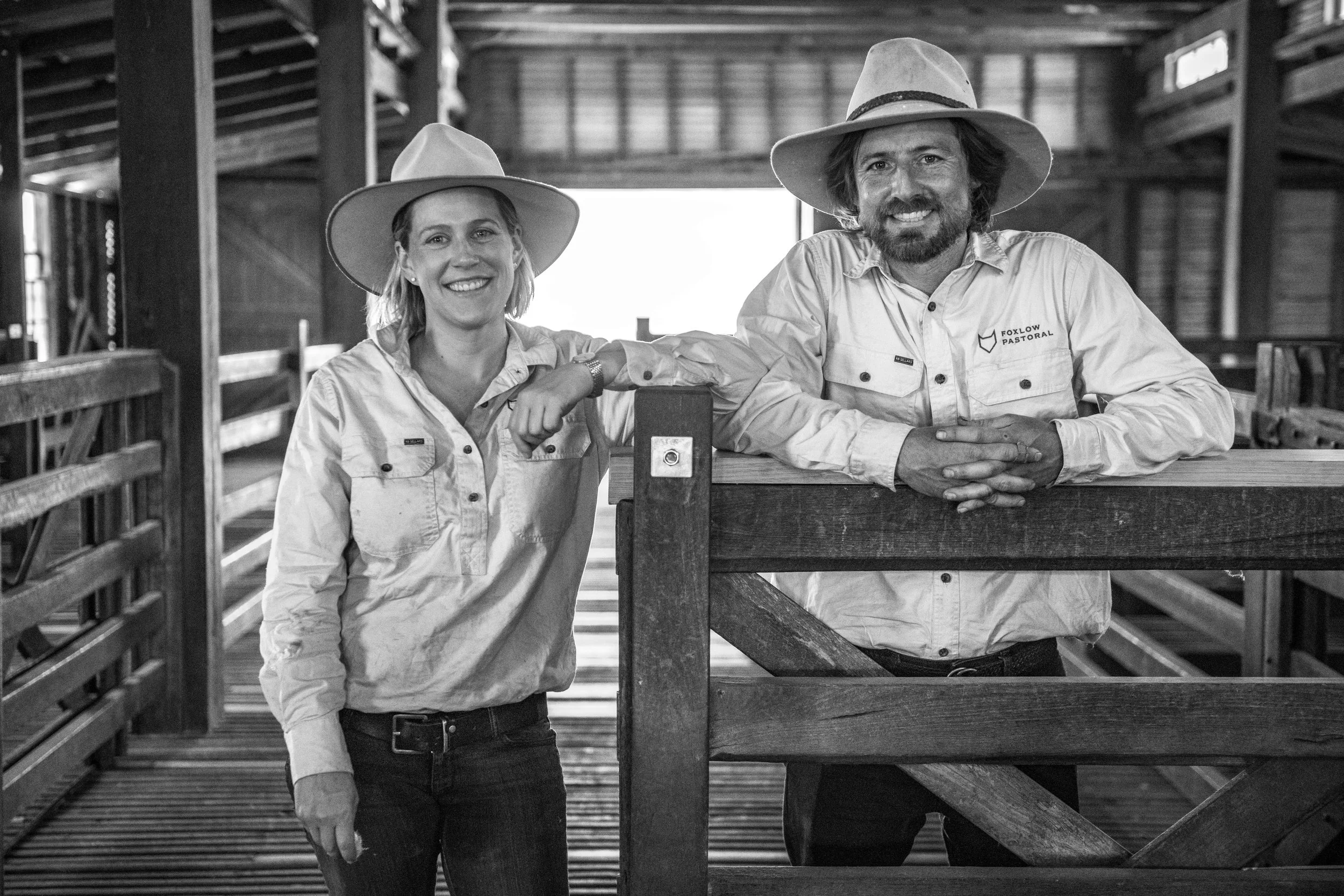 Two smiling farmers, a woman and a man, in cowboy hats and long-sleeve shirts, standing inside a barn with wooden fences, with bright light coming from behind.