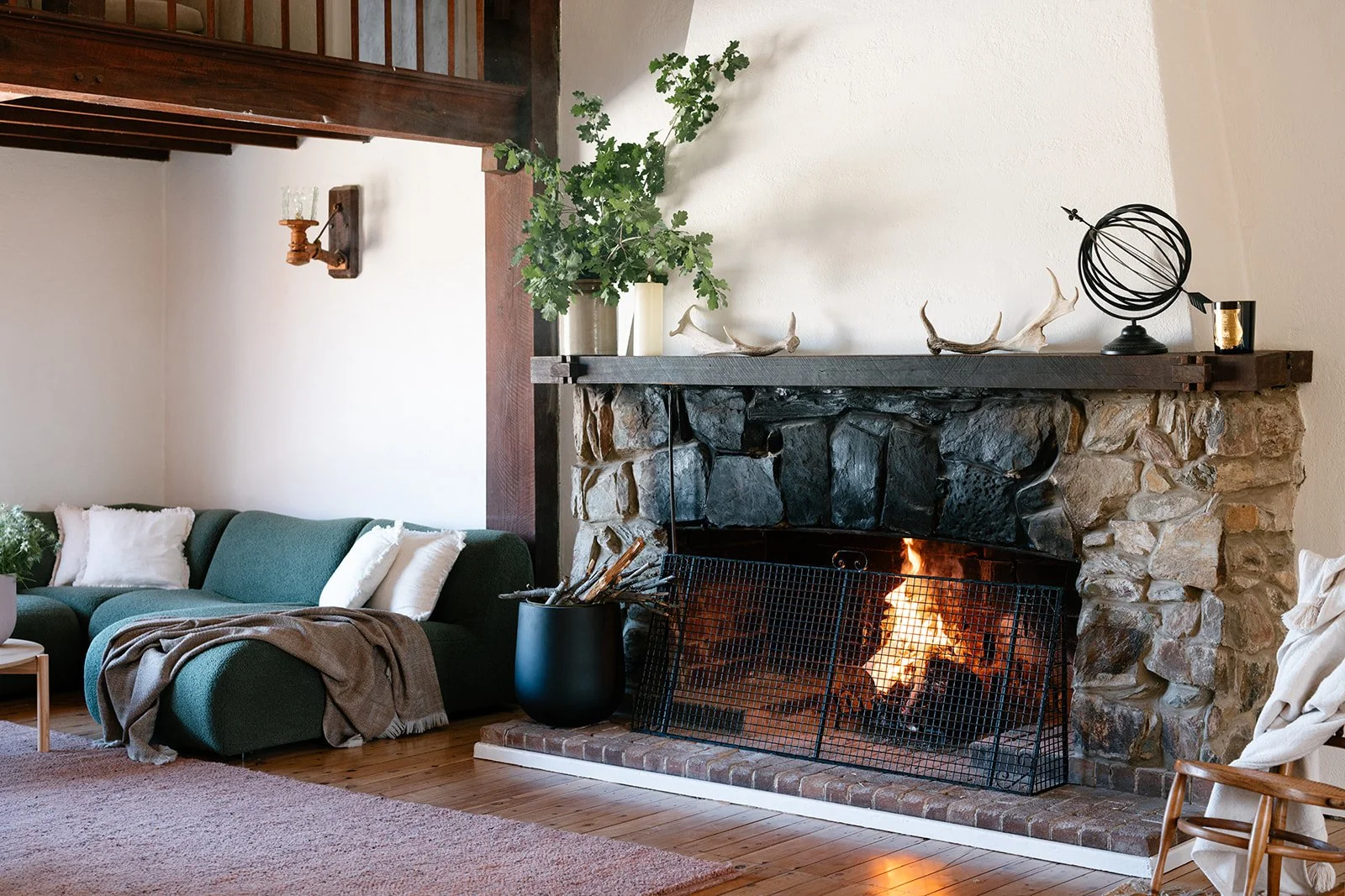 Living room with a stone fireplace, a green sofa with white pillows, a throw blanket, a wooden coffee table, and a wooden chair. Decor items on the mantel include a potted plant, antlers, a spherical sculpture, and a candle. A fire burns in the fireplace.