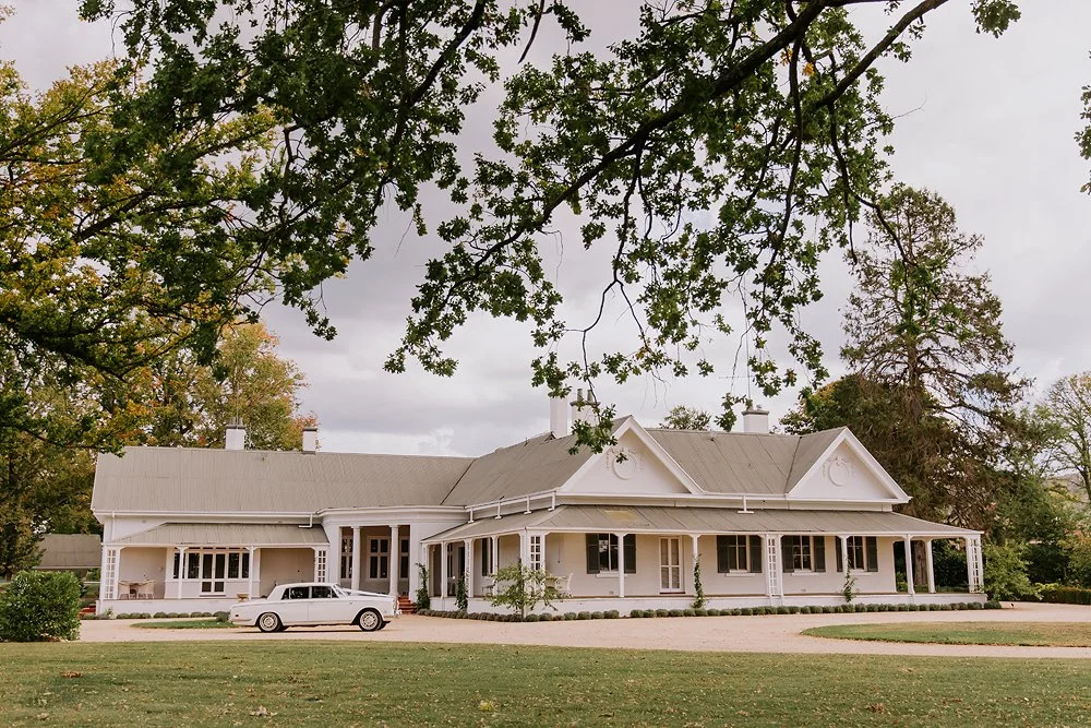 Large white house with a wraparound porch, surrounded by trees and a lawn, parked car in front, and cloudy sky overhead.