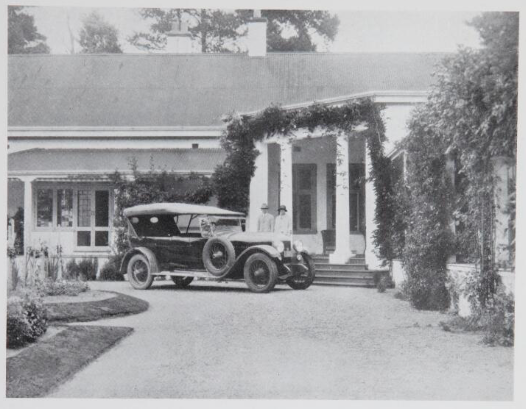 A historic black and white photograph of a vintage car parked in front of a house with a porch, surrounded by greenery and garden plants.