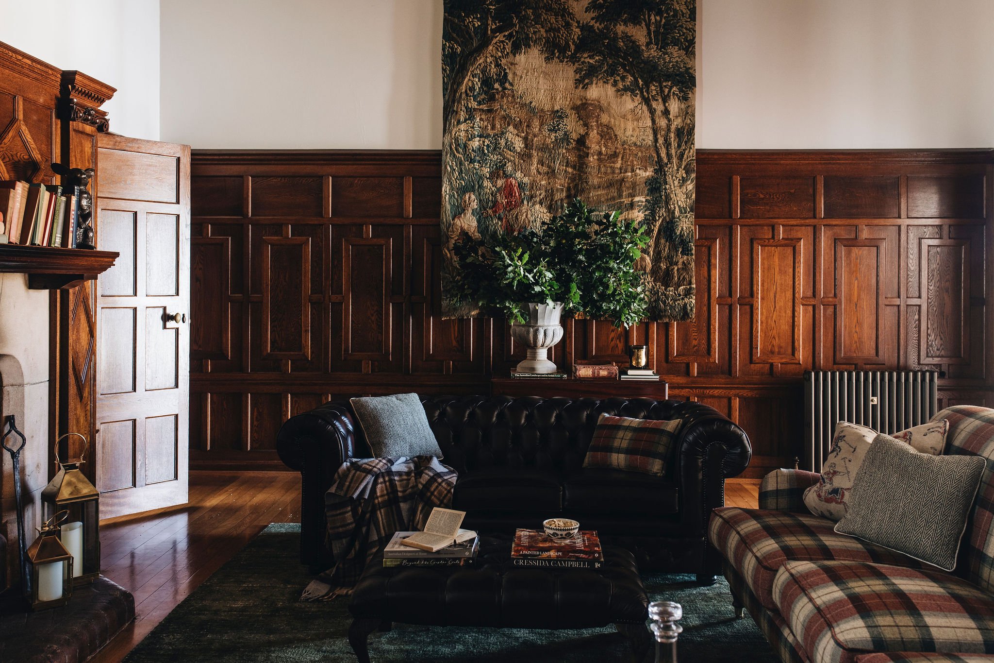A cozy living room with dark wooden paneling on the walls, a tapestry hanging above a large green plant in a white urn, a black leather chesterfield sofa with cushions and a blanket, a plaid sofa with decorative pillows, a stack of books, and a fireplace with candles and lanterns on the hearth.