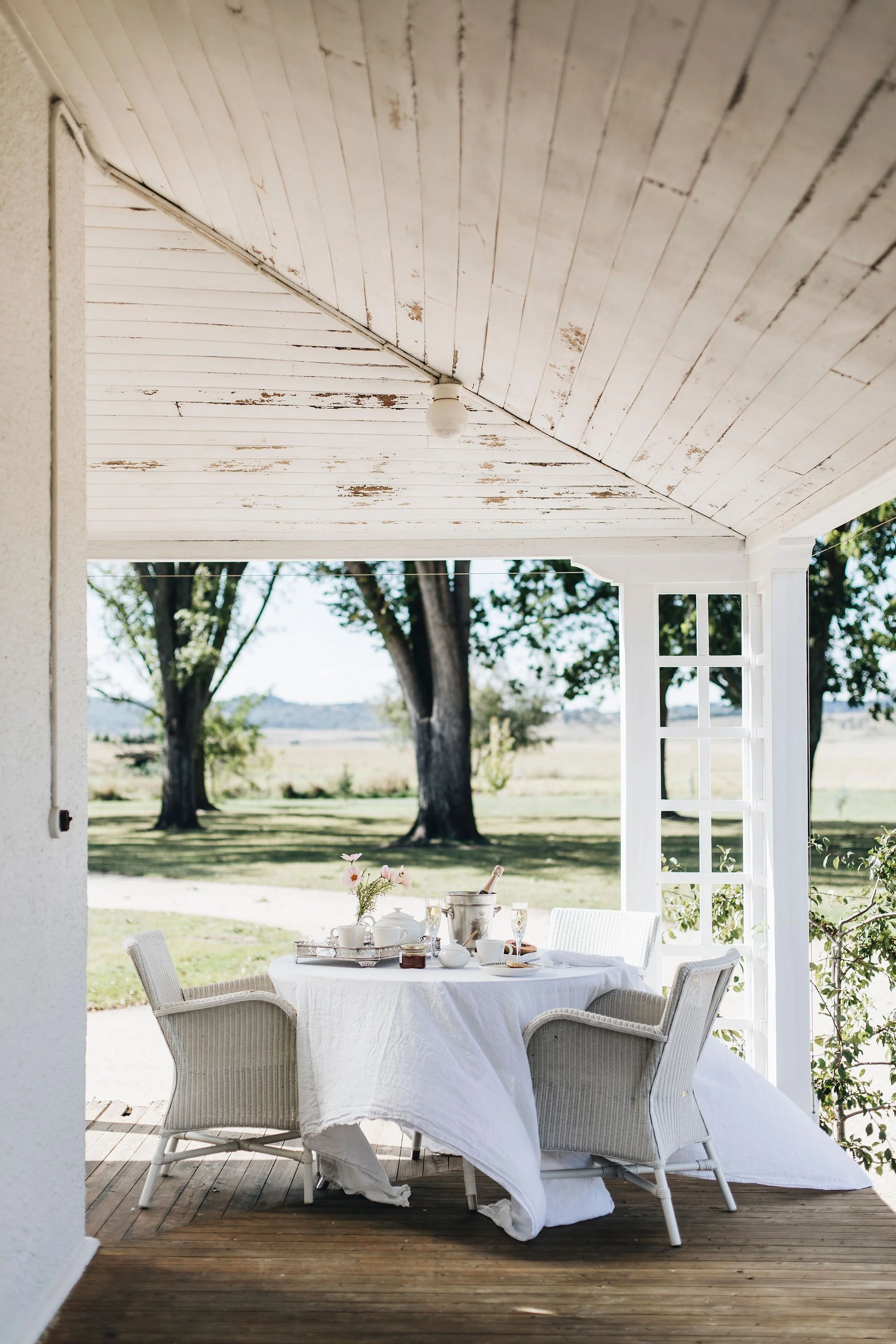 Porch with round table set for tea, four chairs, white tablecloth, flowers, and outdoor scenery.