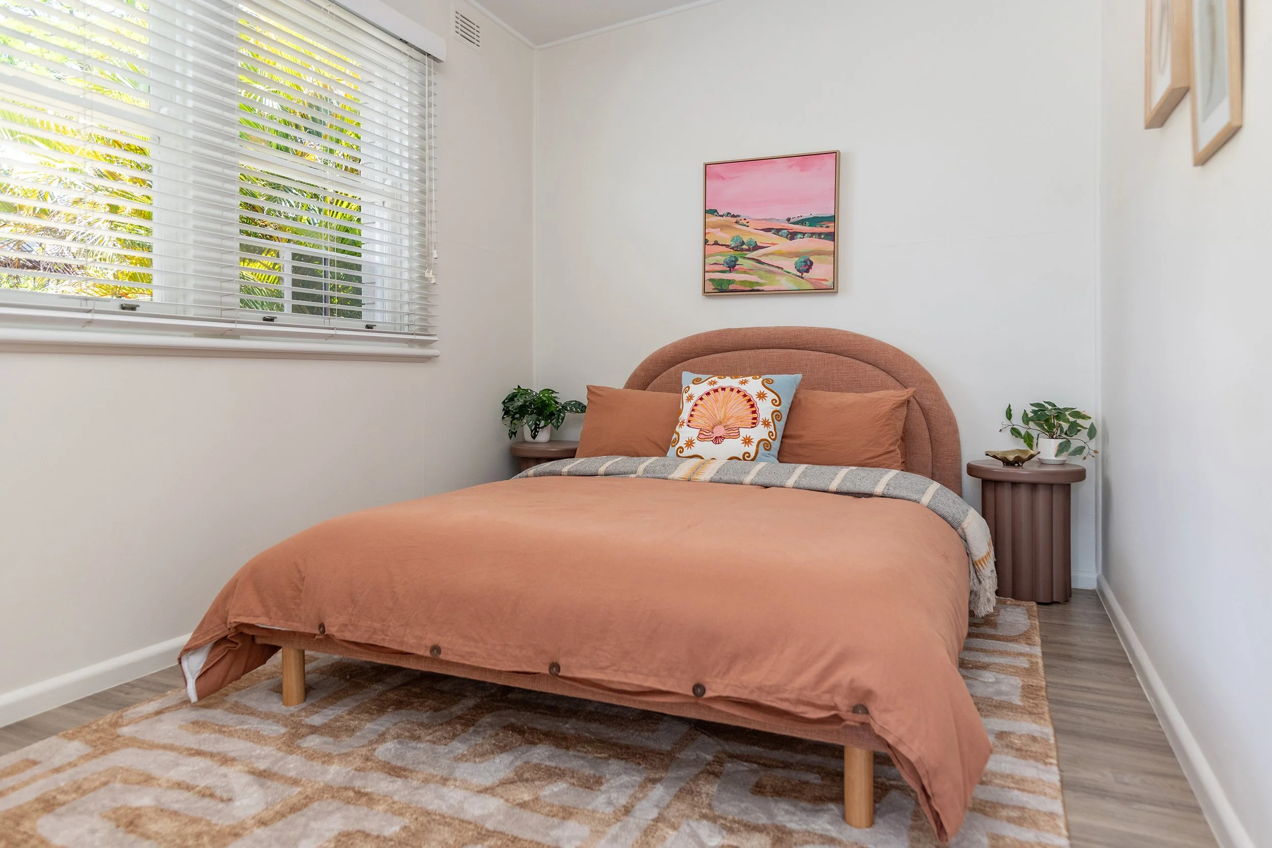 A bedroom with a bed covered in a rust-colored duvet, two matching pillows, decorative pillow with a seashell design, bedside tables with potted plants, window with white blinds, and a colorful landscape painting on the wall.
