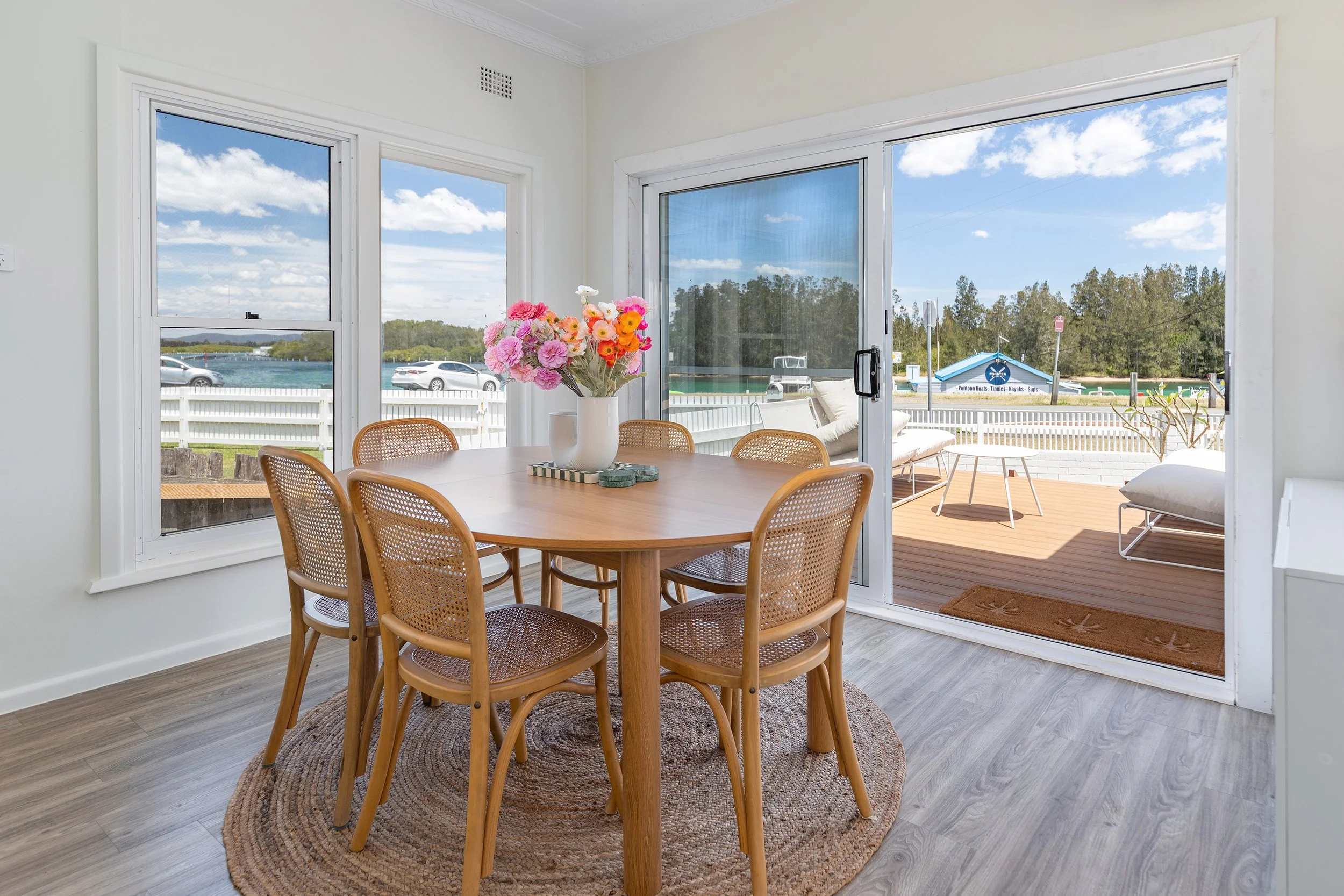 Dining area with a wooden table and six wicker chairs, a vase of colorful flowers, and large windows with a view of an outdoor patio, water, boats, and cars.