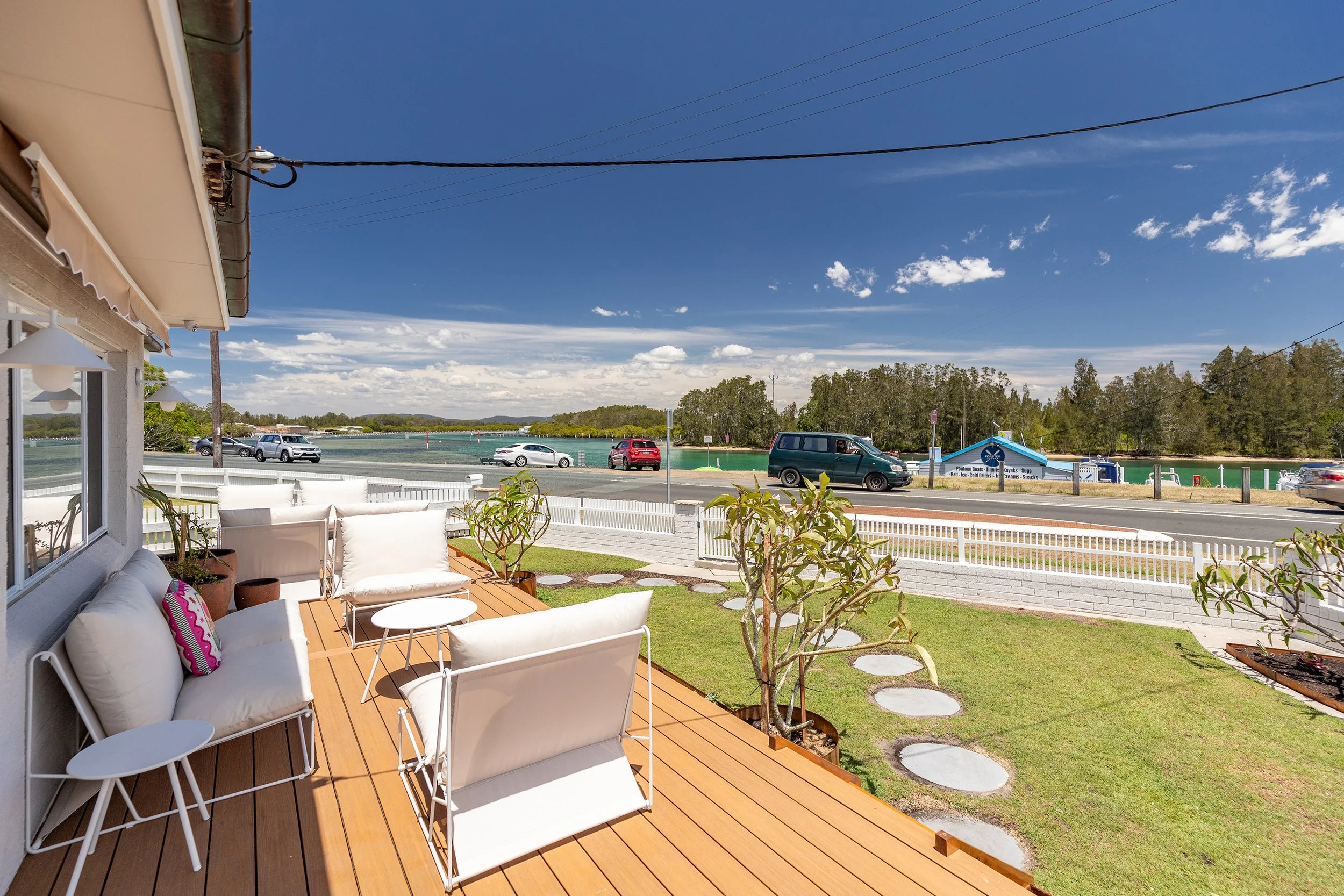 A wooden porch with white outdoor furniture and potted plants overlooking a waterfront with cars parked along the street, trees, and a blue sky with scattered clouds.
