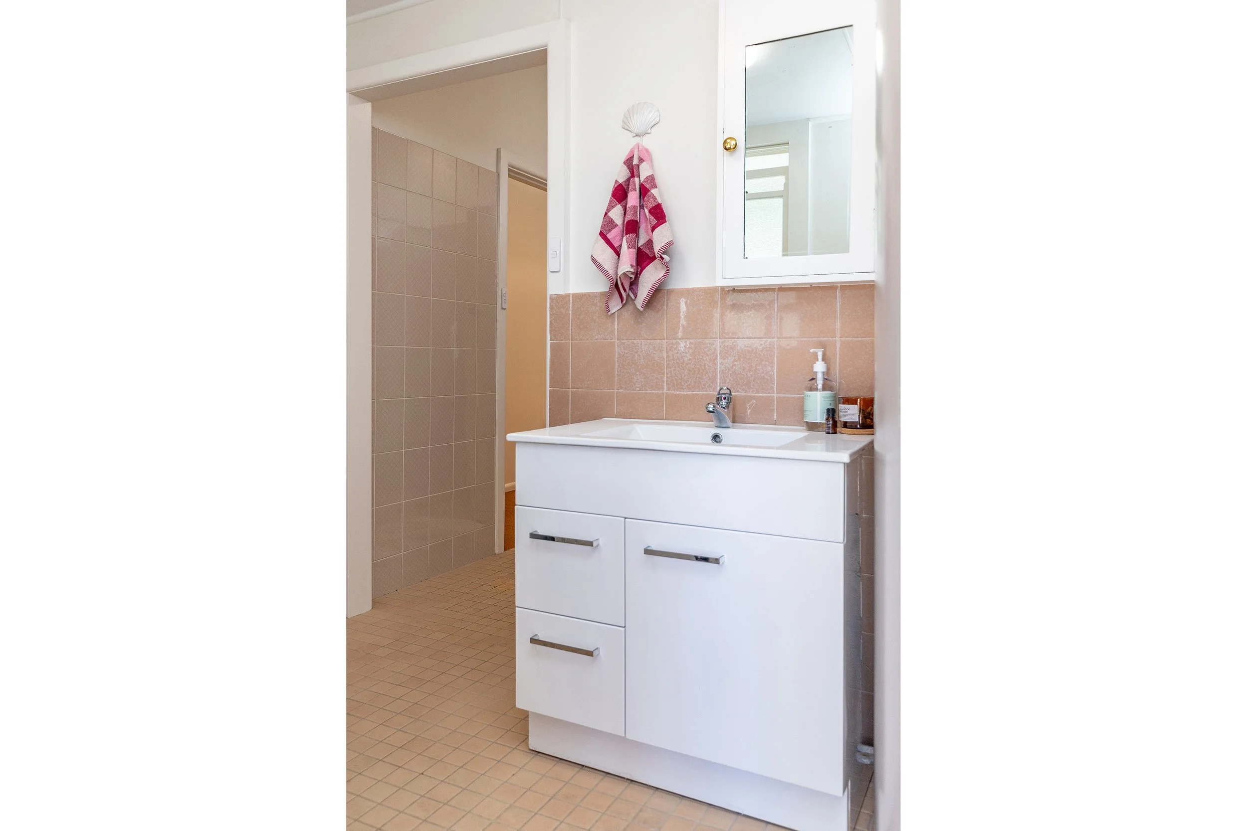 Bathroom with white vanity, pink tile walls, a mirror cabinet, a towel hanging on a shell-shaped hook, bottle of liquid soap, and a small container on the sink, with a shower area visible in the background.