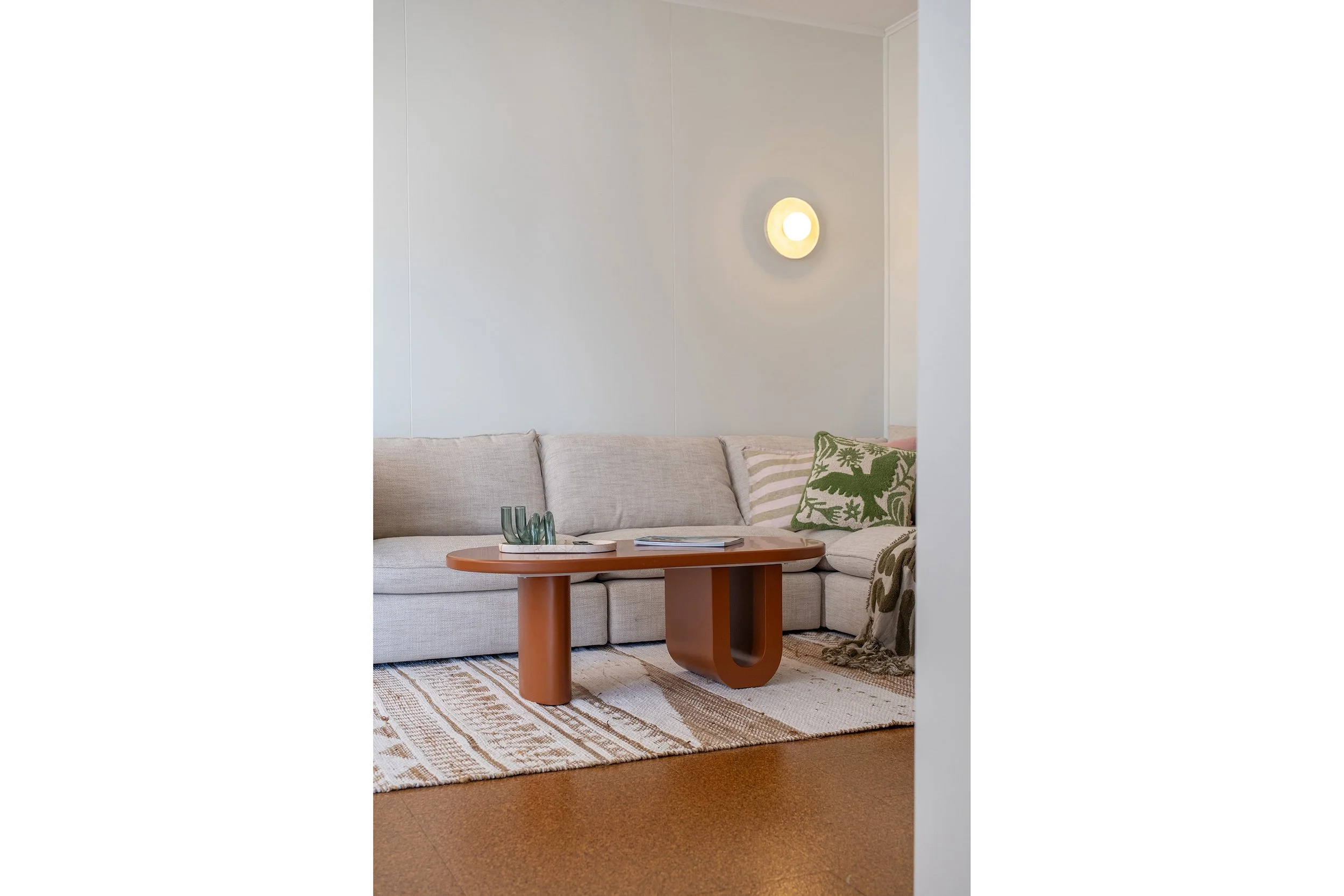 Living room with a beige sofa, patterned pillows, a wooden coffee table, decorative glass pieces, and a wall-mounted light fixture.