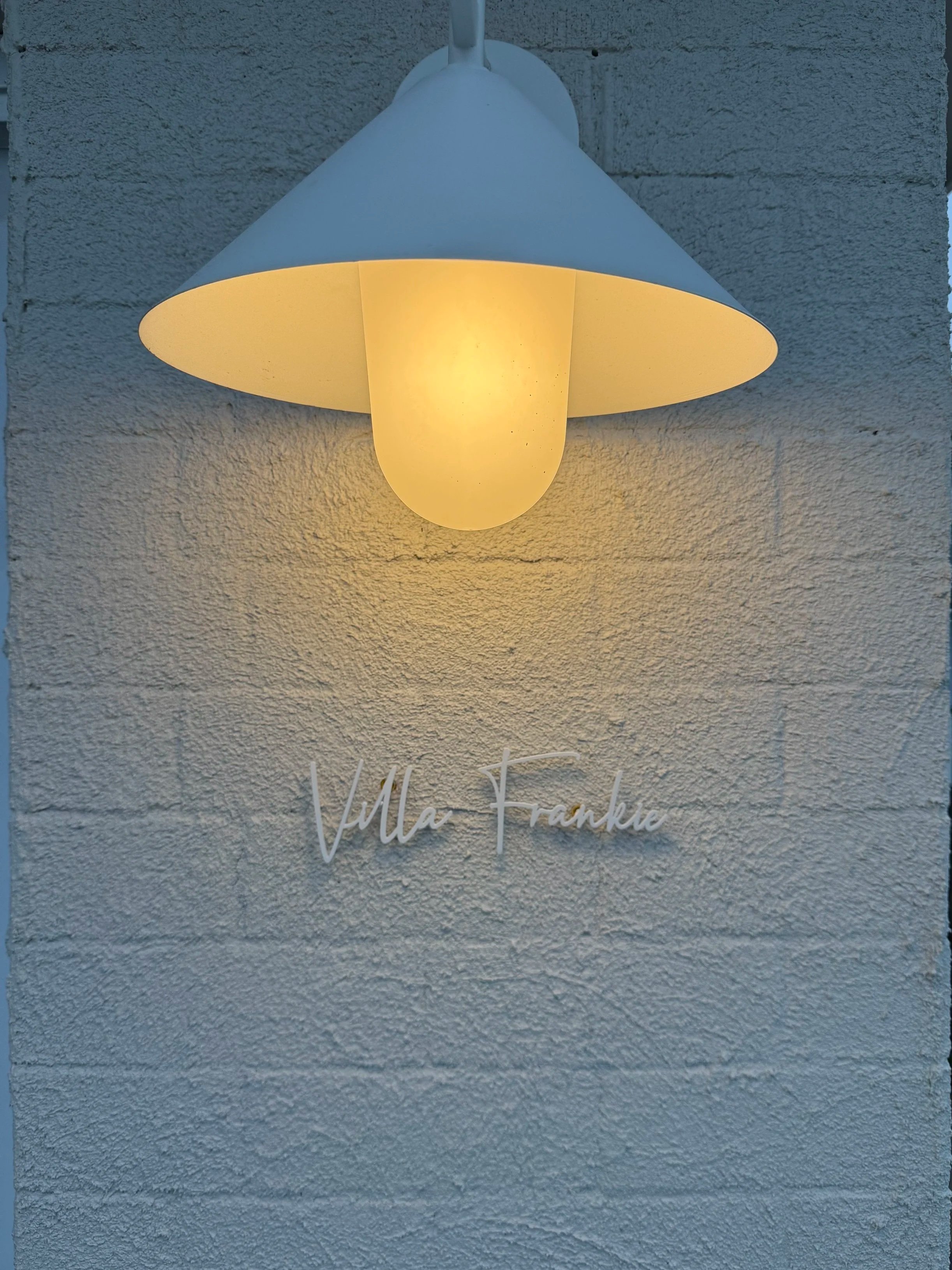 Wall-mounted lamp with a white shade and warm light bulb, with 'Villa Frankie' written in cursive on textured white wall.