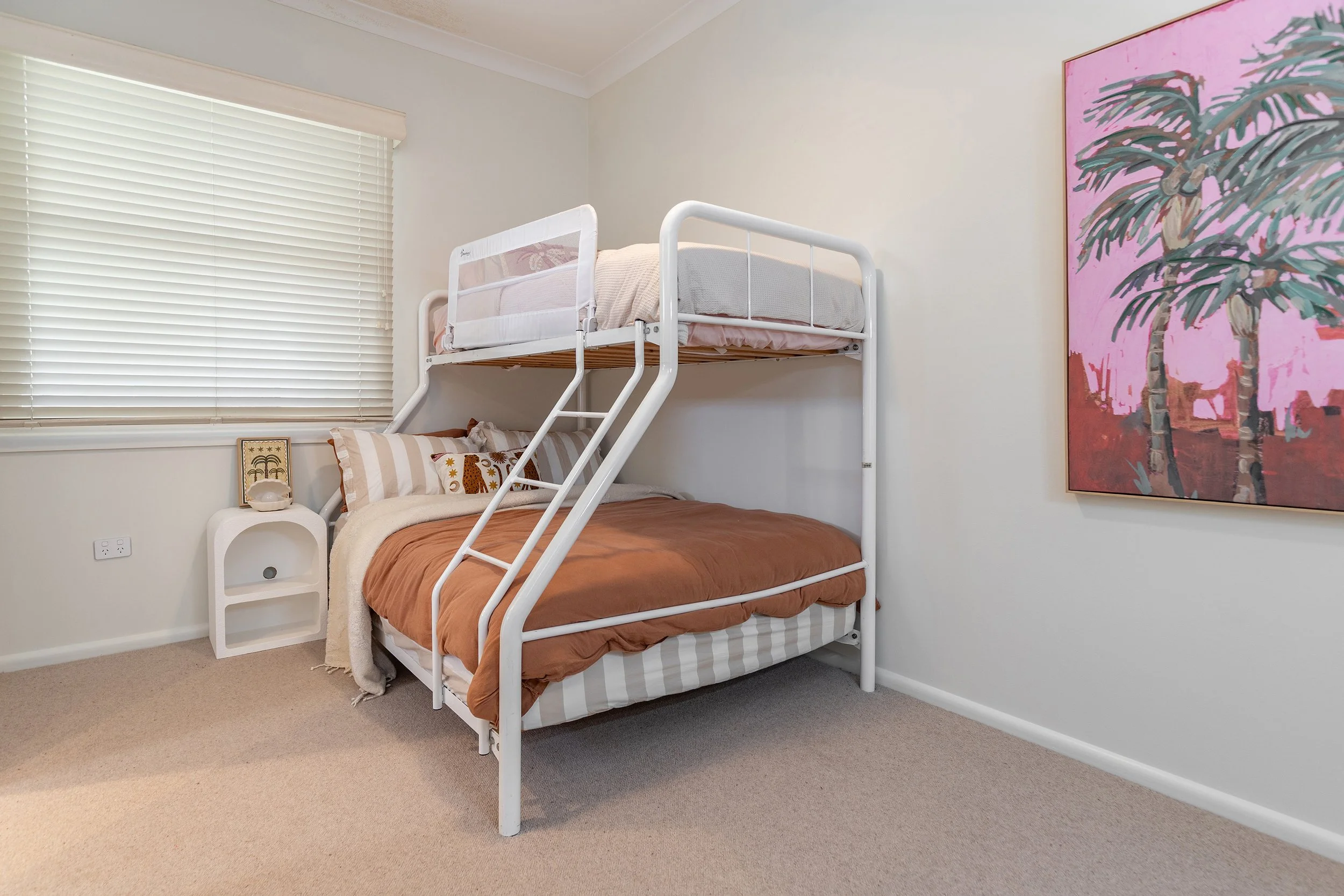 A bedroom with a white bunk bed, brown bedding on the lower bunk, and a white bedrail on the upper bunk. A window with closed blinds is on the left, and a pink palm tree painting is on the right wall.