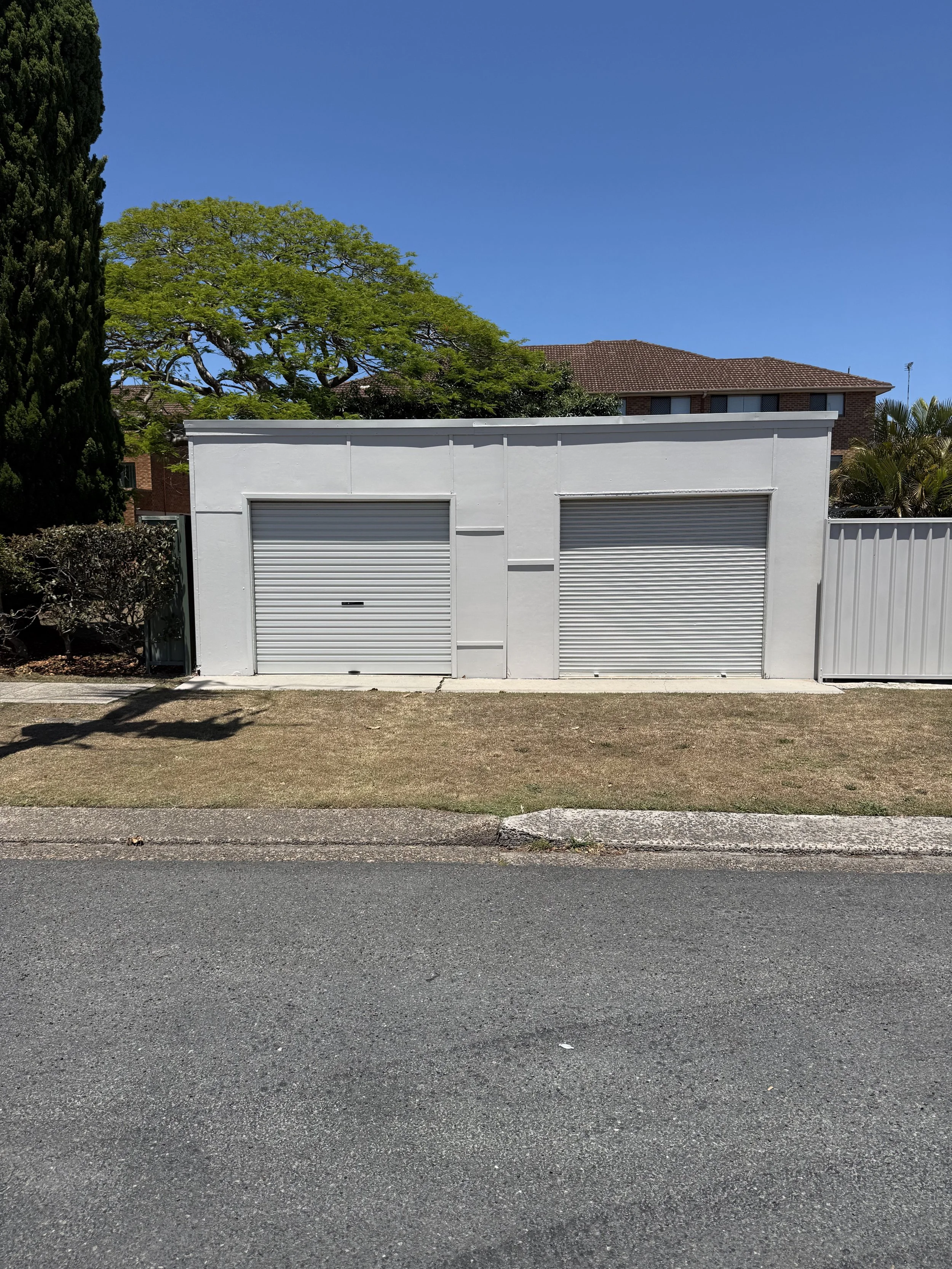 A small white building with two closed roller doors, situated on a sidewalk with a grassy area in front, under a clear blue sky.