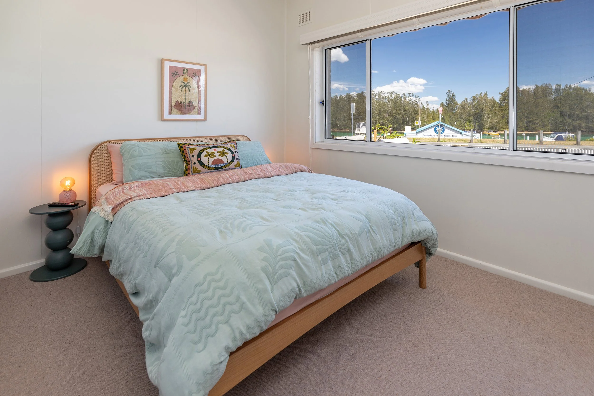 Bedroom with a bed, colorful pillows, a nightstand with a lamp, and a large window showing a blue sky and outdoor scene.