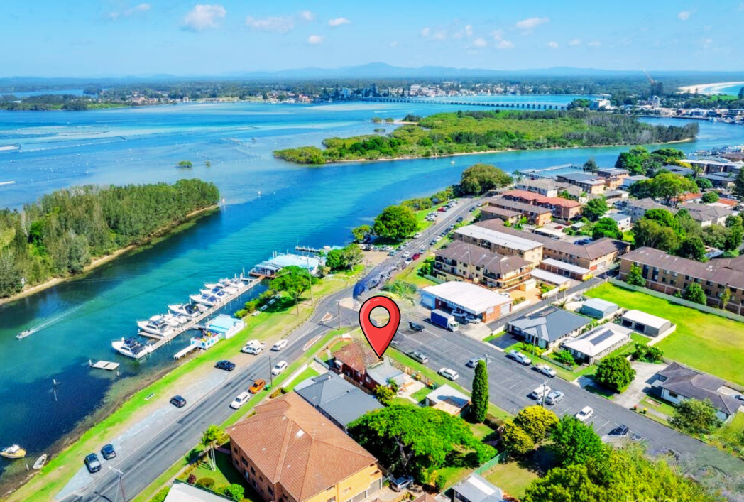 Aerial view of a lakeside neighborhood with boats docked at a marina, surrounding greenery, residential buildings, and a large body of water with islands and a distant bridge.