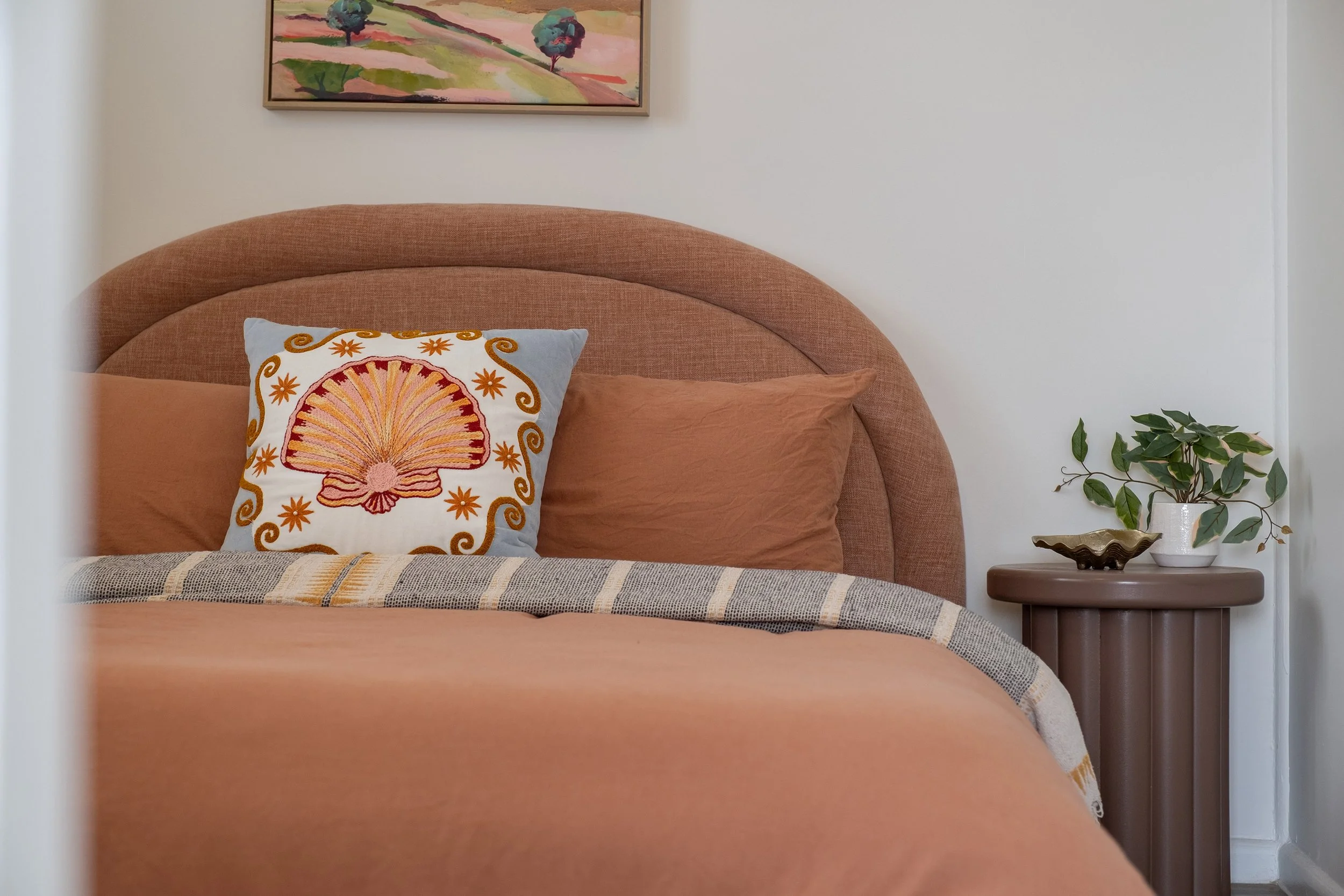 A bedroom with a peach-colored bedspread, matching pillows, and a decorative pillow with an embroidered shell and starfish design. A framed landscape painting hangs above the bed. To the right, there is a round, brown bedside table with a potted plan