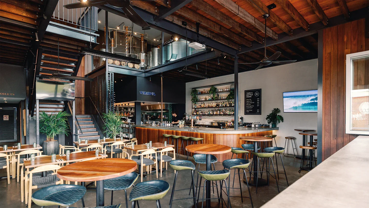 Interior of a modern bar and restaurant with wooden furniture, a bar counter, and a staircase leading to the upper level.