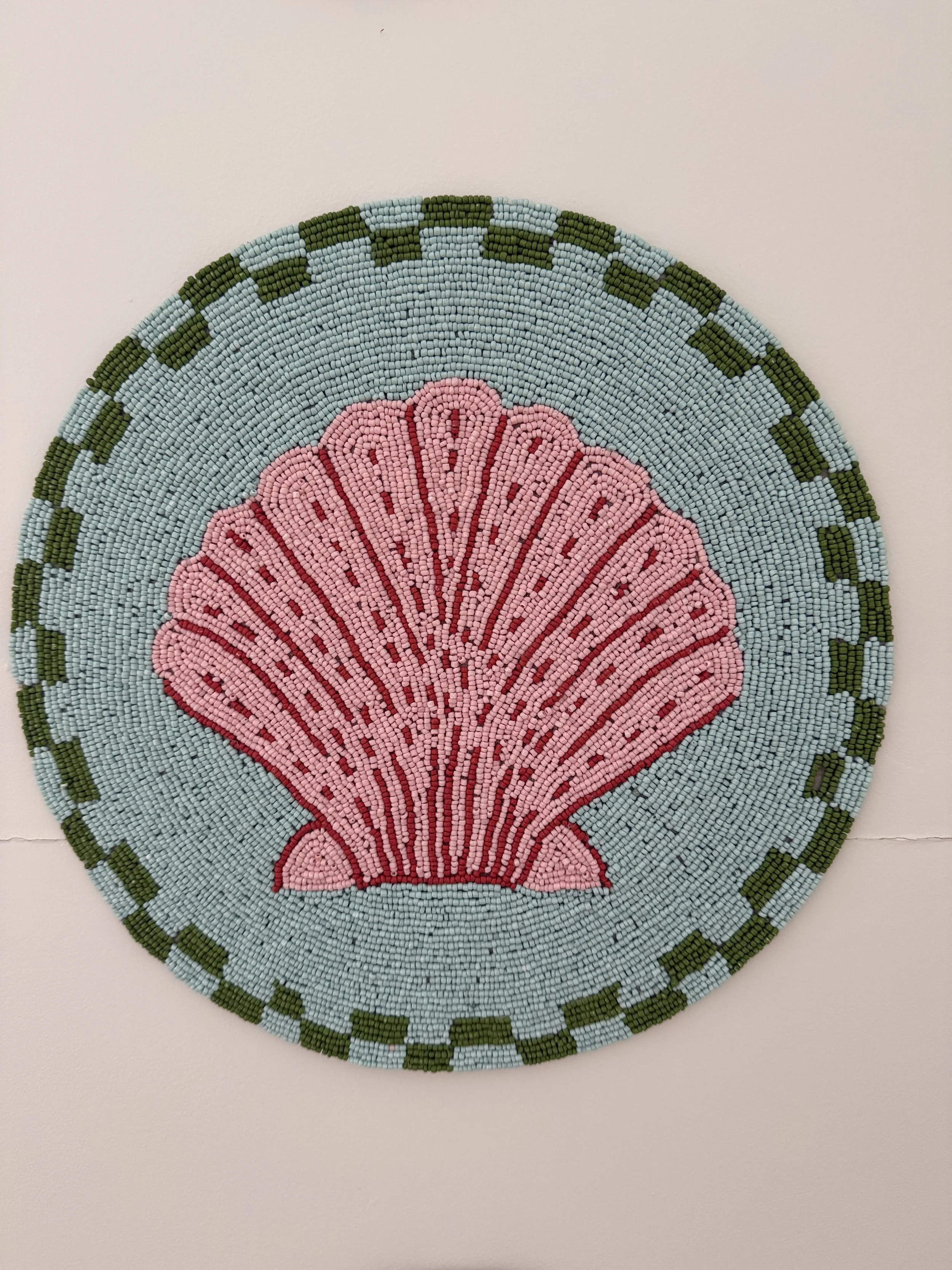 Beaded round wall art featuring a pink seashell on a light blue background with a green and dark green checkered border.