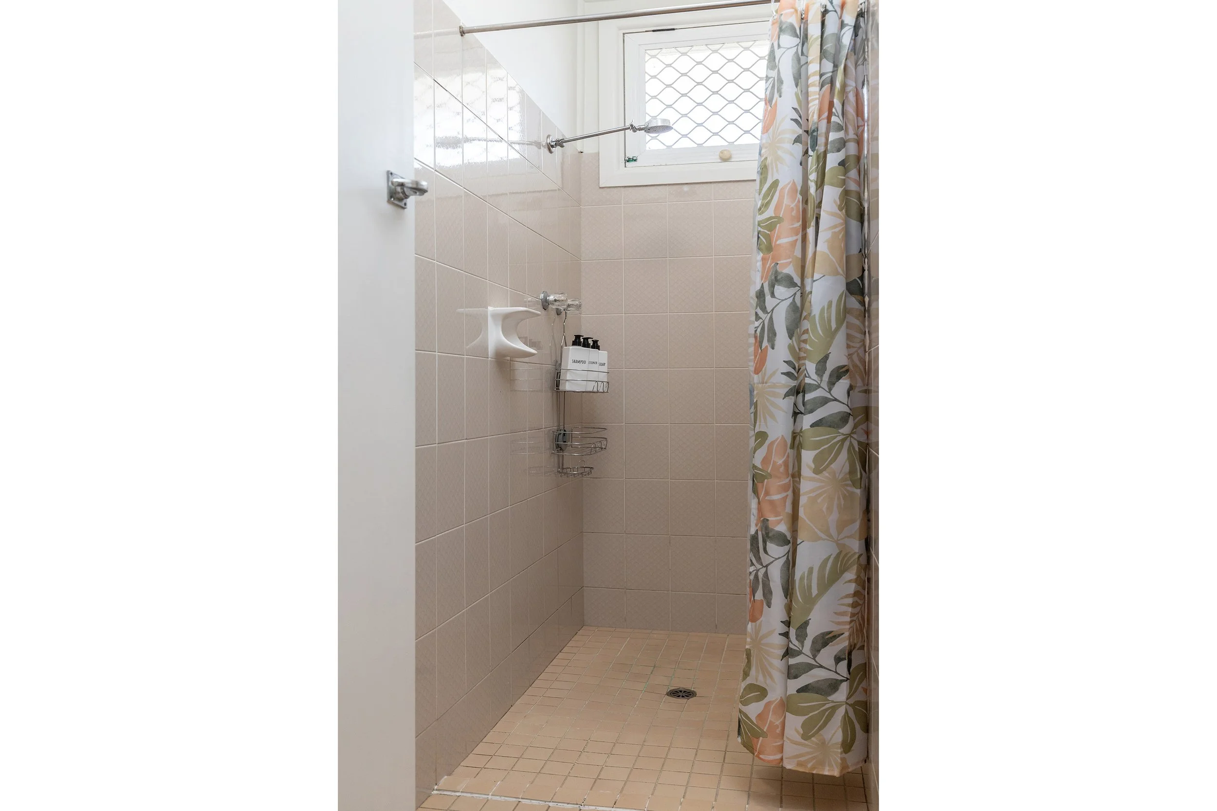 Empty shower with beige tiled walls and floor, a window at the top, a floral shower curtain, a white soap dish, and shelves with shampoo bottles.
