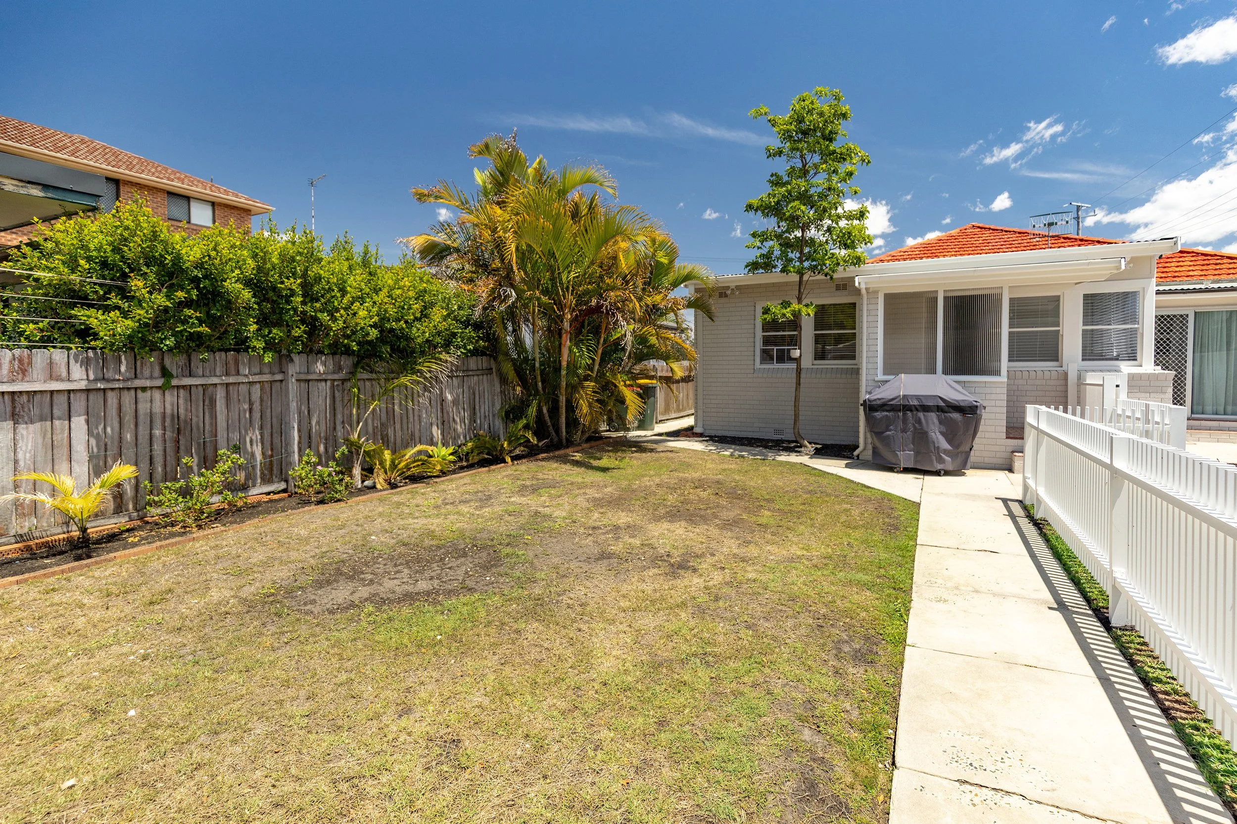 Backyard yard with patchy grass, a concrete pathway, white fence, small trees, bushes, a covered barbecue grill, and a house with a brick exterior and a red tile roof under a blue sky.