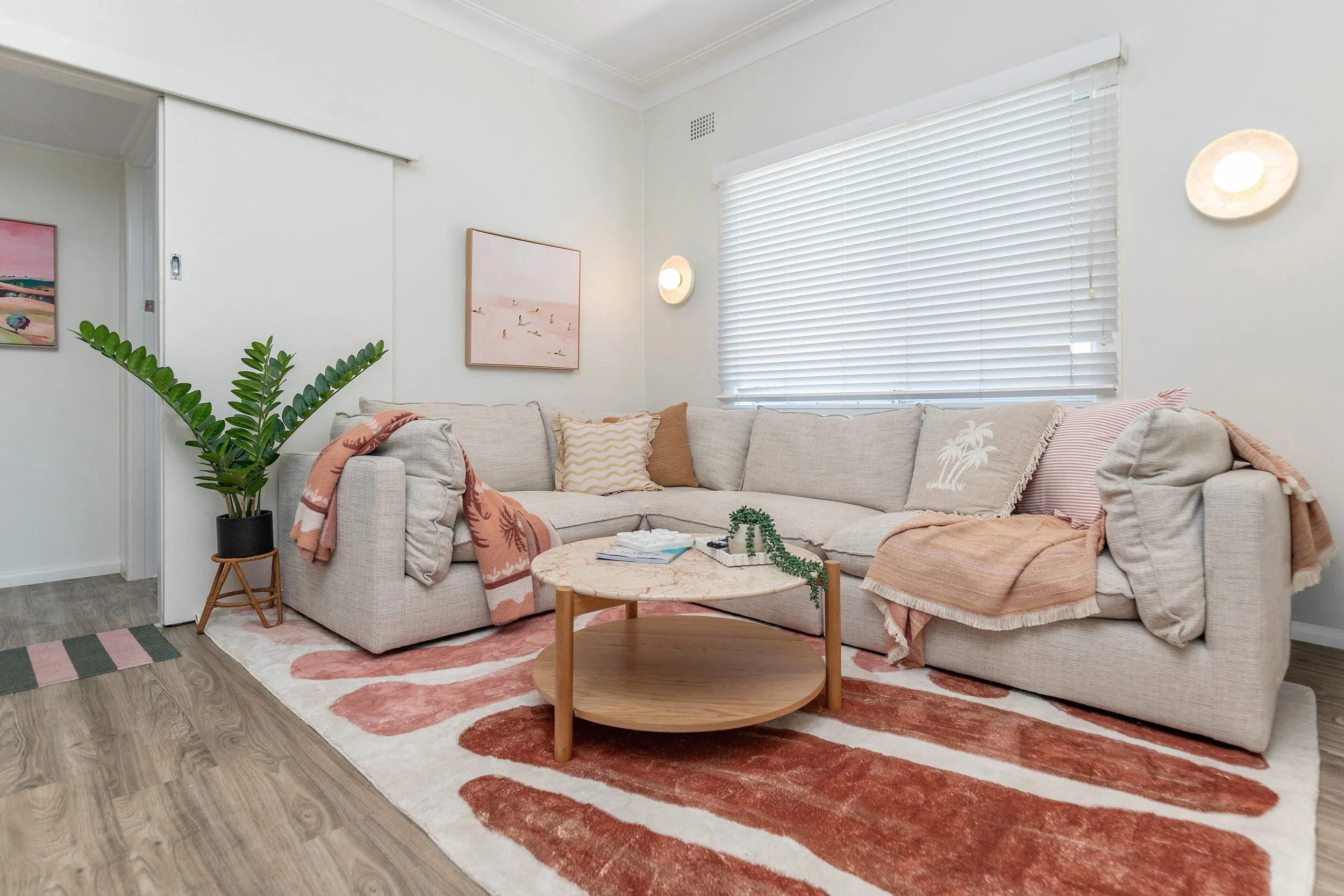 A cozy living room with a beige sectional sofa decorated with pastel and patterned cushions, a round marble-top coffee table with a plant and books, a large window with white blinds, a potted plant in a black pot, wall-mounted light fixtures, and art