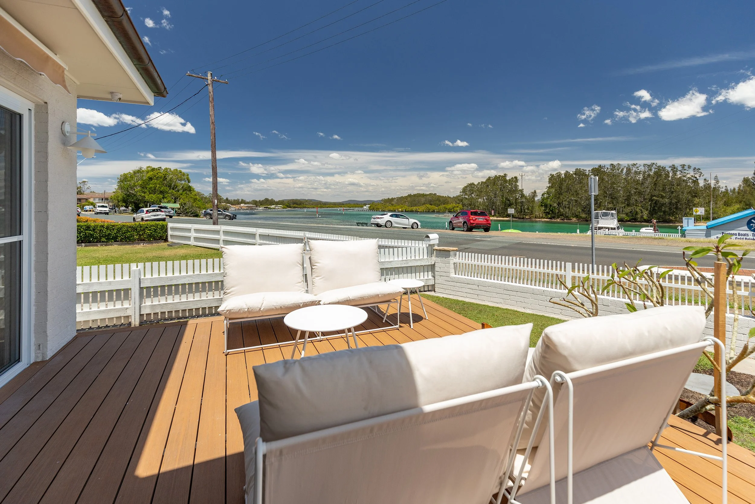 A wooden outdoor deck with white cushions and small white tables, overlooking a road, a waterway, and trees under a partly cloudy blue sky.