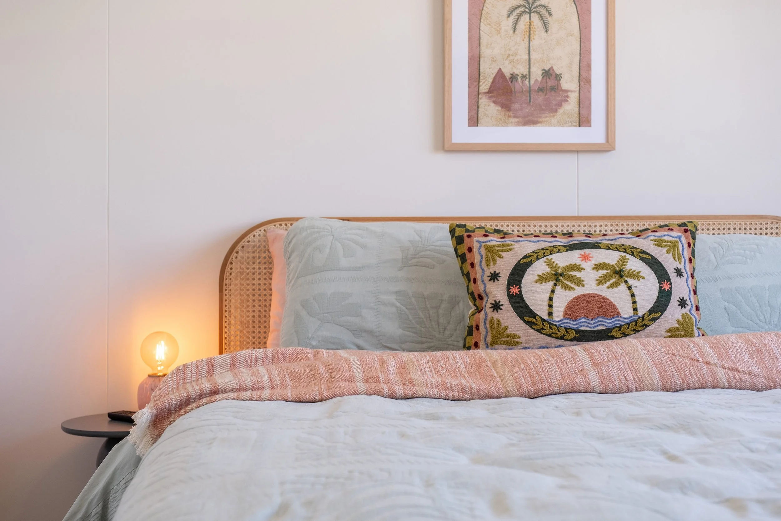 A neatly made bed with a light-colored quilt, a decorative pillow featuring palm trees and a sunset, and a pink strip of fabric at the bottom. There is a bedside table with a small lamp casting a warm glow, and a framed picture of a palm tree on a beach hangs on the white wall above the bed.