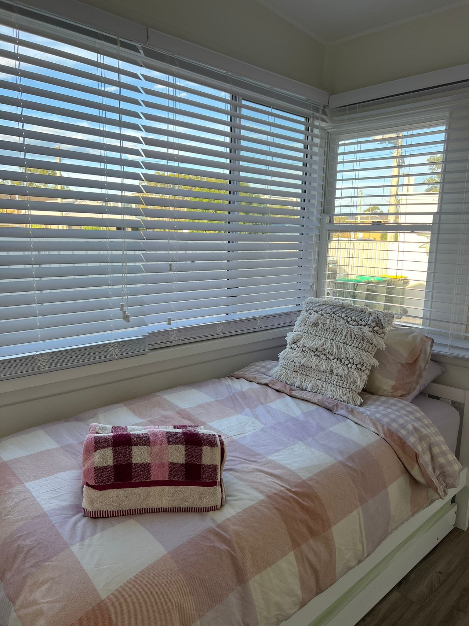 A bed with a plaid blanket, a decorative pillow, and a folded towel on top, situated near large windows with white blinds, in a bright room with natural sunlight.