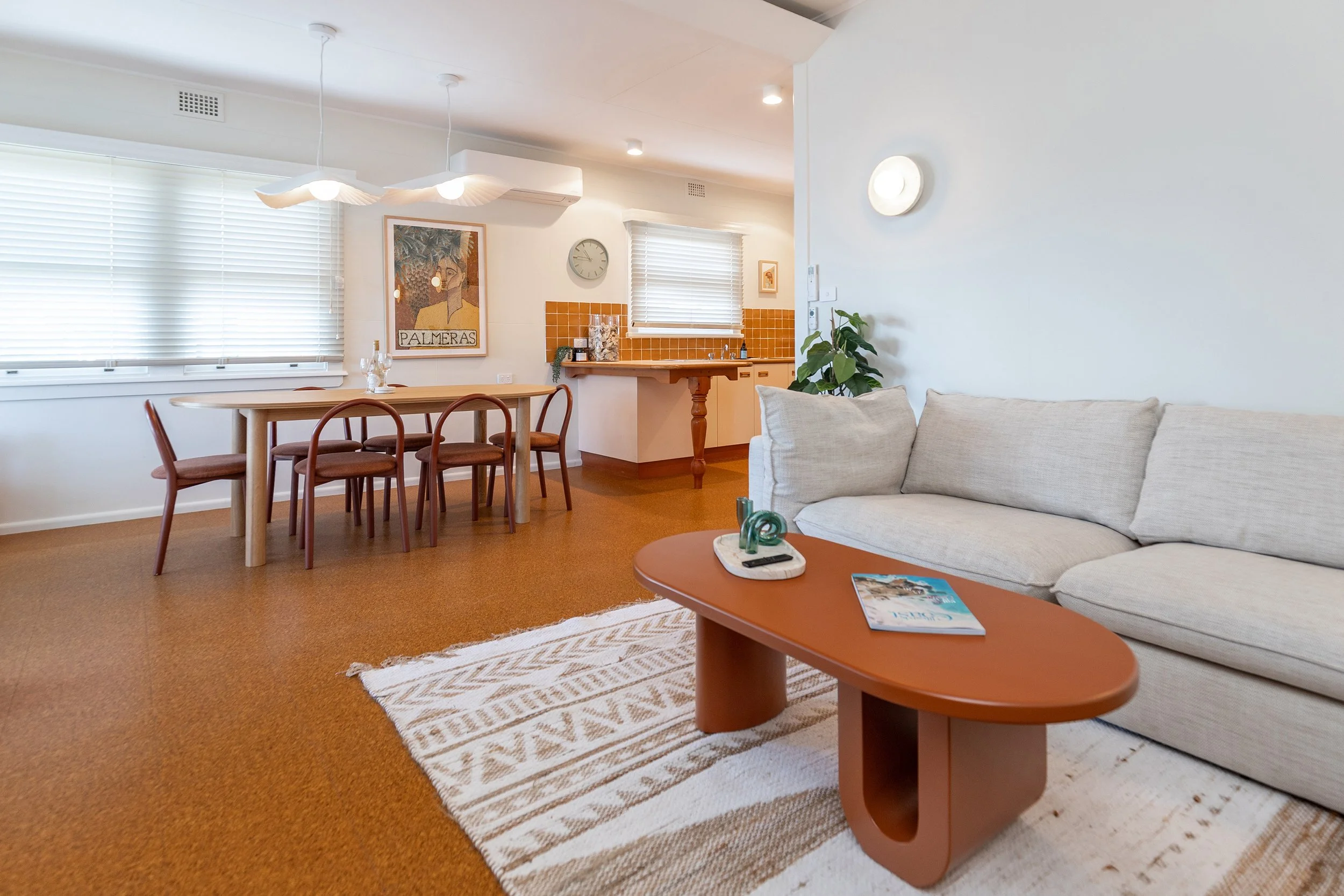 Living room and dining area with a white sofa, wooden coffee table with decorative items and magazine, rug, and a dining table with six chairs, kitchen counter with orange tiles, windows with blinds, wall art, and a clock.