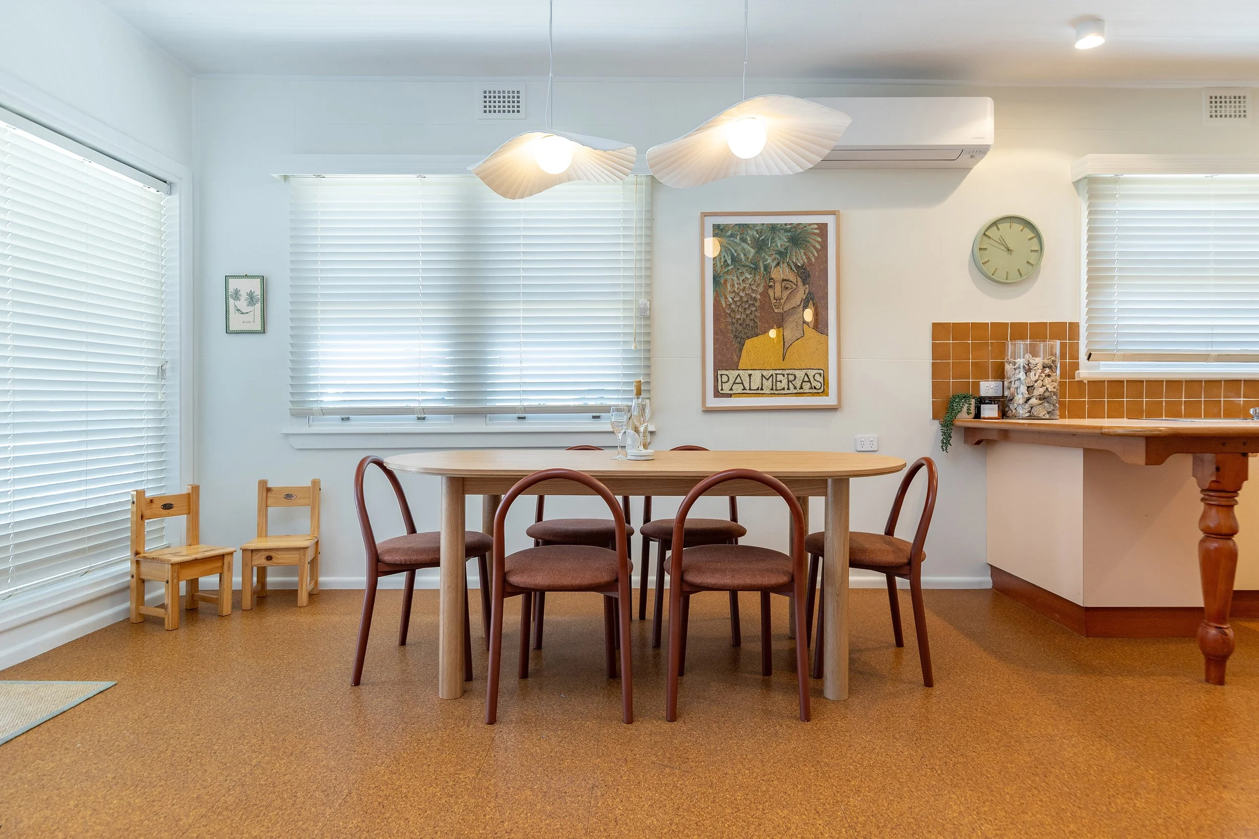 Dining room with a wooden table and six chairs, white window blinds, framed artwork on the wall, a clock, and a side counter with a glass jar and some greenery.