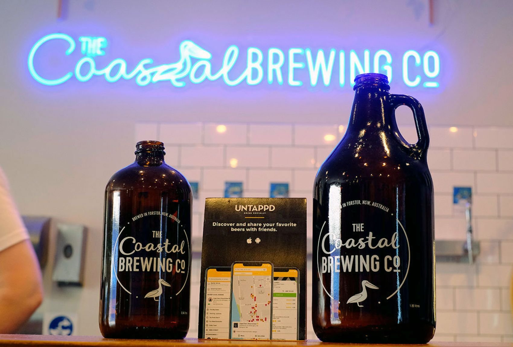 Two brown glass bottles with The Coastal Brewing Co. logo placed on a counter, with a promotional sign and a neon sign that says 'The Coastal Brewing Co.' in the background.