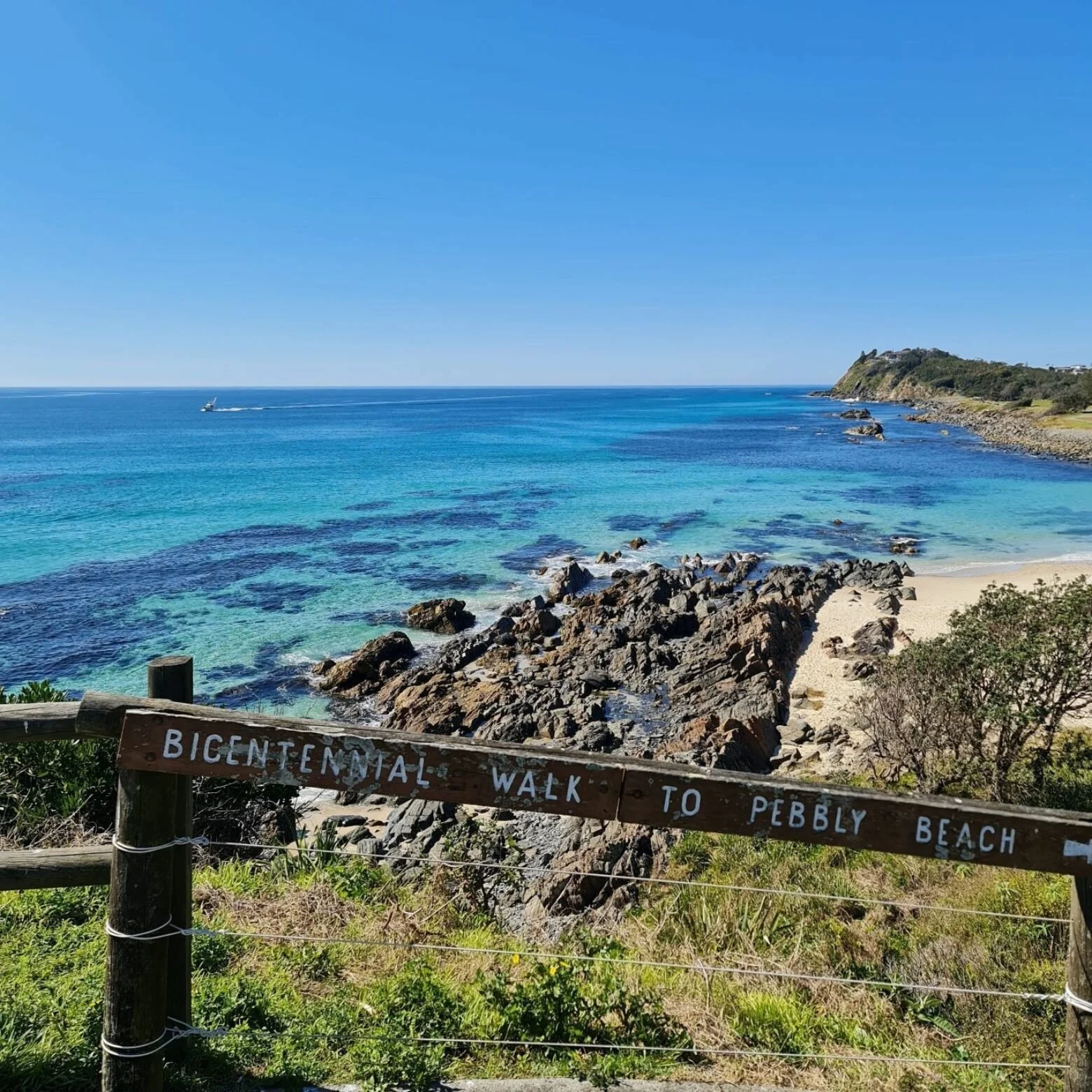 A scenic coastal view of Pebbly Beach with rocky shoreline, turquoise water, and cliffs in the distance. There is a wooden sign in the foreground indicating the Bicentennial Walk to Pebbly Beach.