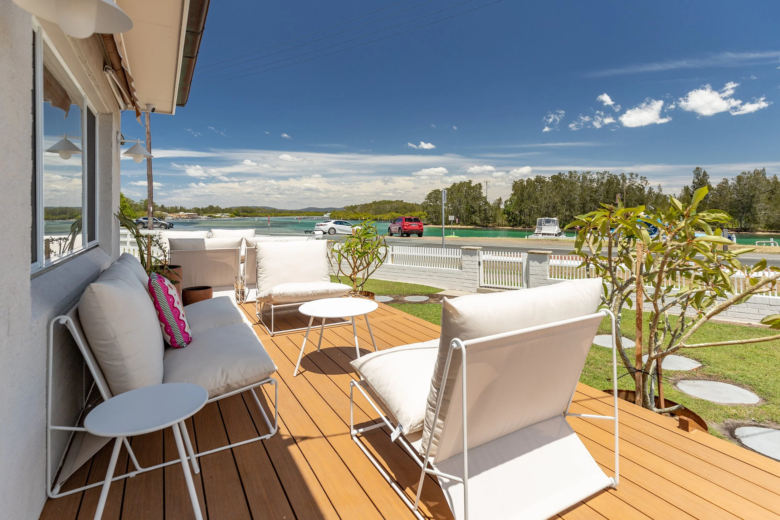 View of a patio with white cushioned outdoor furniture, small white tables, and potted plants on a wooden deck, overlooking a waterway with boats and cars parked nearby under a partly cloudy sky.