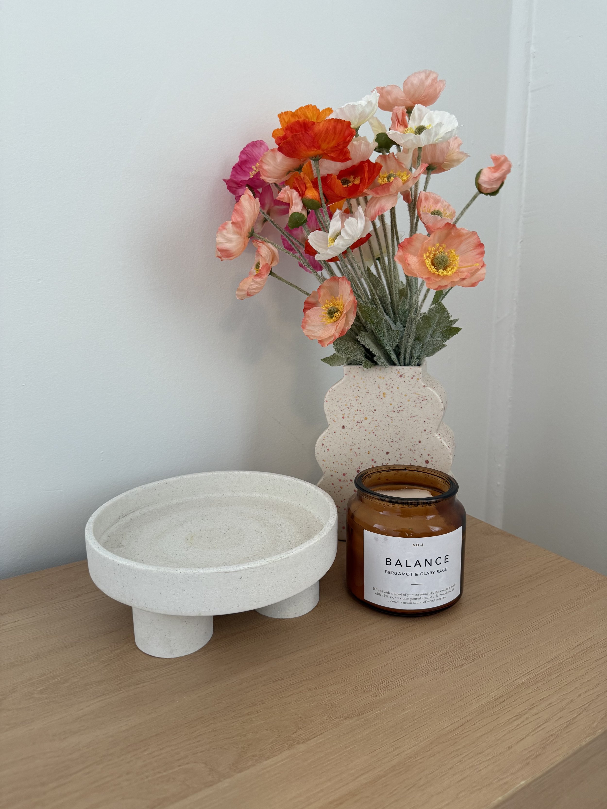 A bouquet of pink, white, orange, and red flowers in a white speckled vase, placed on a wooden surface with an empty white speckled dish and a brown candle labeled 'Balance' with bergamot and clary sage scent.