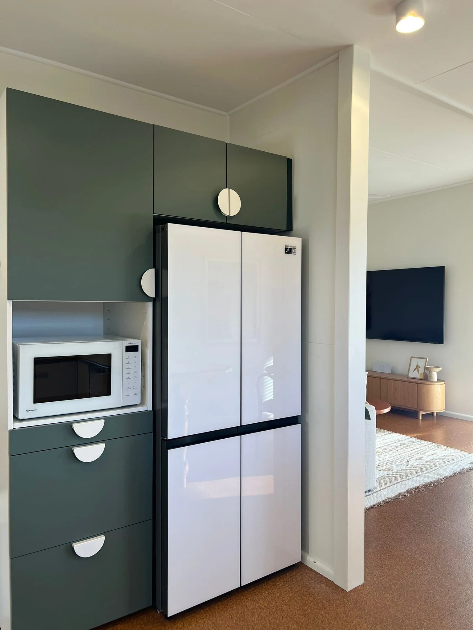 A kitchen area with green cabinets, a white microwave on a shelf, and a white refrigerator with dual doors. In the background, a living room with a flat screen TV, wooden cabinet, and patterned area rug.