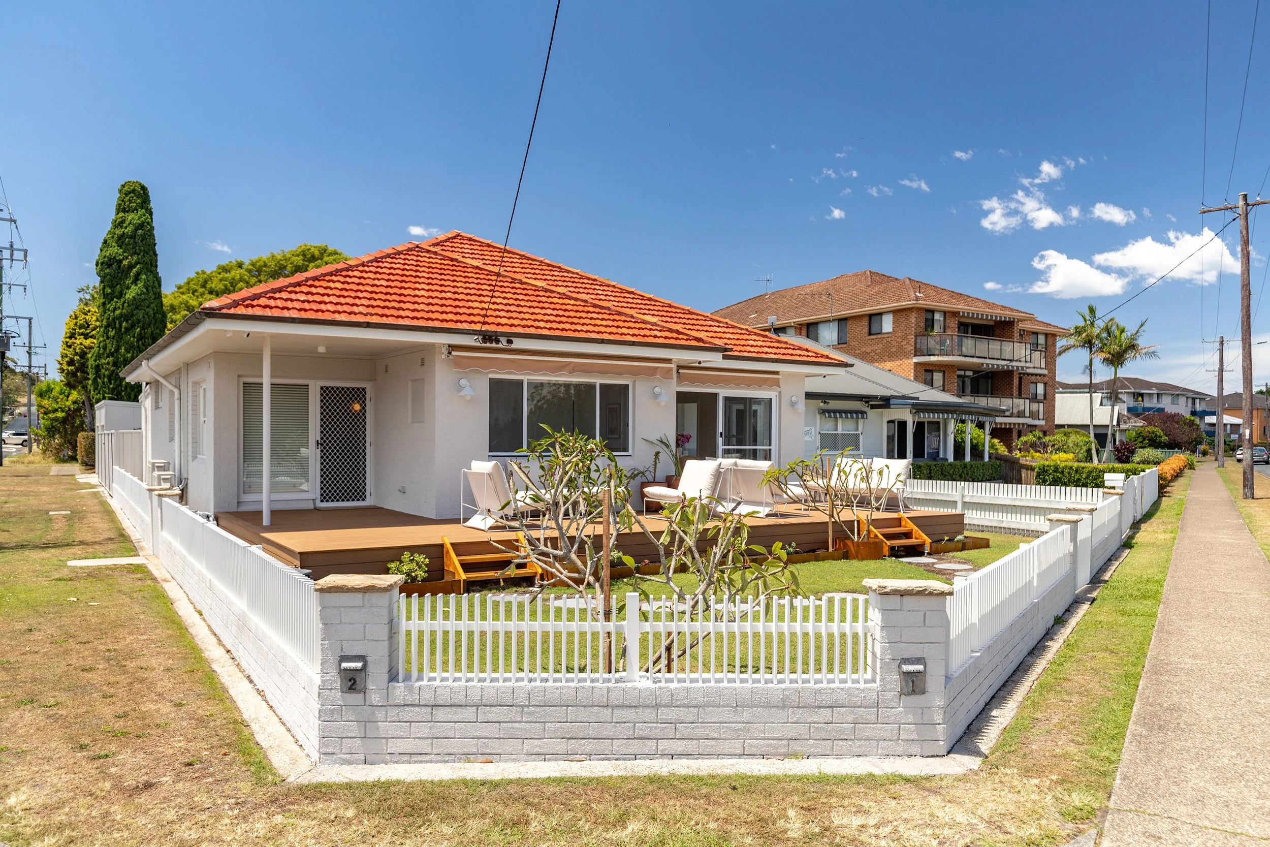 A white house with a red tiled roof surrounds a front yard with a wooden deck, white chairs, and a small tree, all enclosed by a white picket fence on a sunny day.