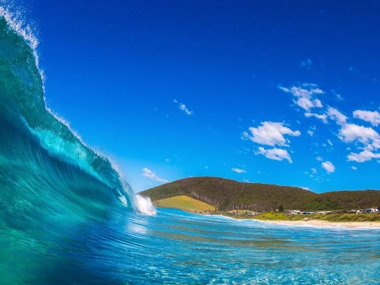 A large ocean wave crashing toward the shore with a green hillside and blue sky in the background