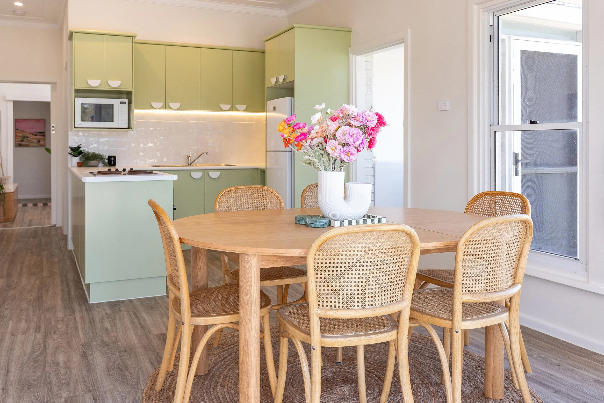Dining room with wooden table, six woven rattan chairs, vase with pink and white flowers, green kitchen cabinets, and white walls.