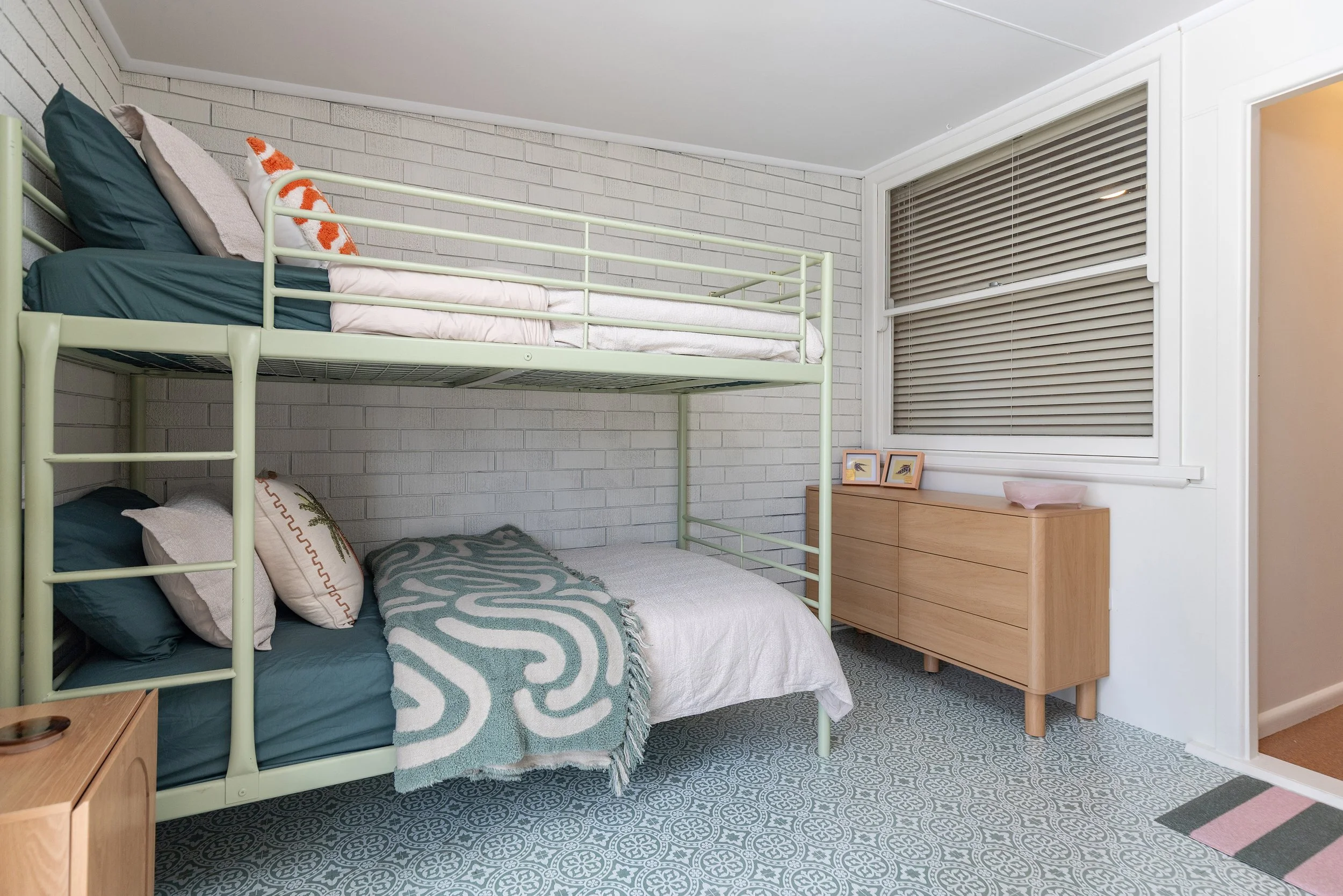 Bedroom with a white brick wall, a bunk bed with green metal frame and dark teal bedding, a wooden dresser, framed pictures, and a window with closed blinds.