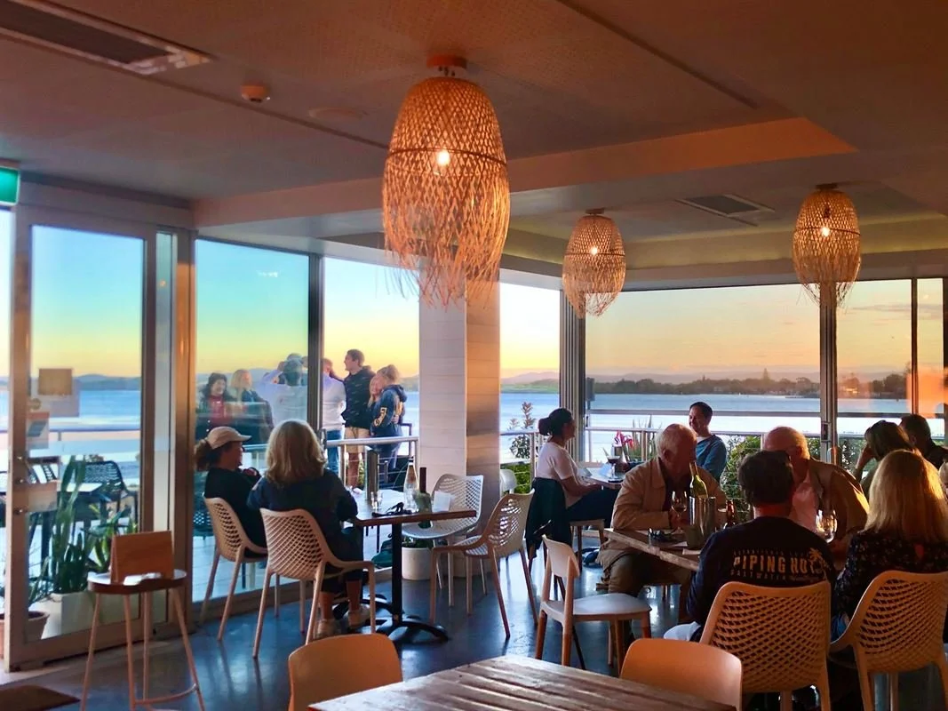 People dining inside a restaurant with large windows overlooking a sunset over water and a deck outside with additional people