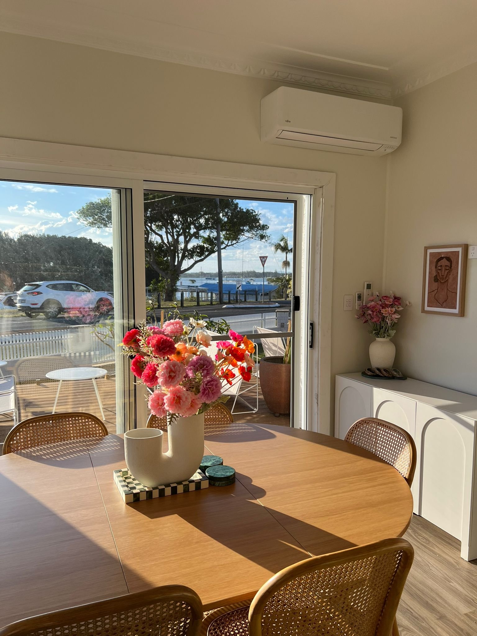 A dining area with a round wooden table decorated with a vase filled with pink and red flowers, located next to a sliding glass door. Outside, there is a patio, a tree, and a view of a marina with boats, bright blue sky, and some clouds.
