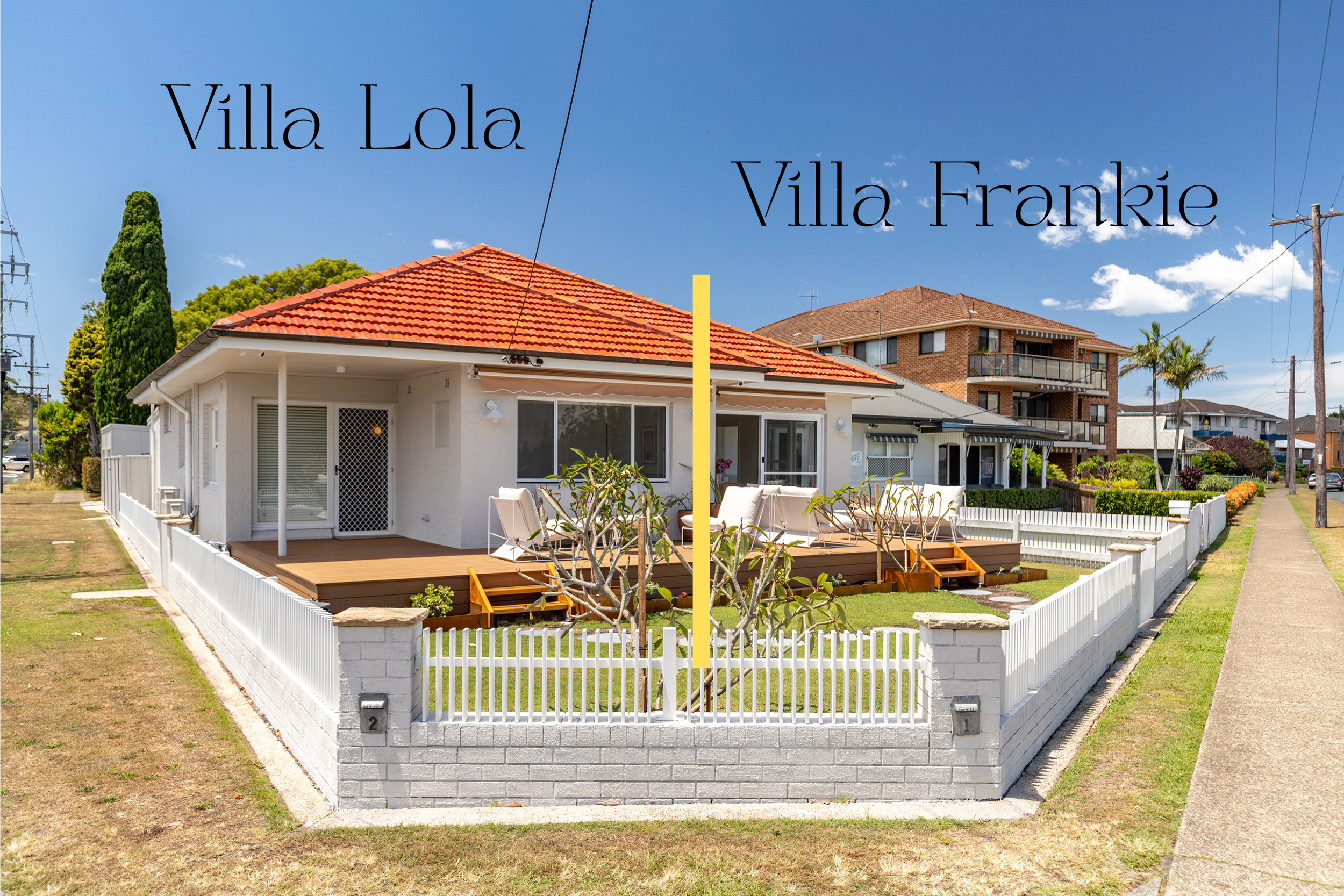 A duplex house with a white exterior and a red-tiled roof, divided into two villas labeled Villa Lola and Villa Frankie, with a small fenced yard and a wooden deck in front, under a blue sky with a few clouds.