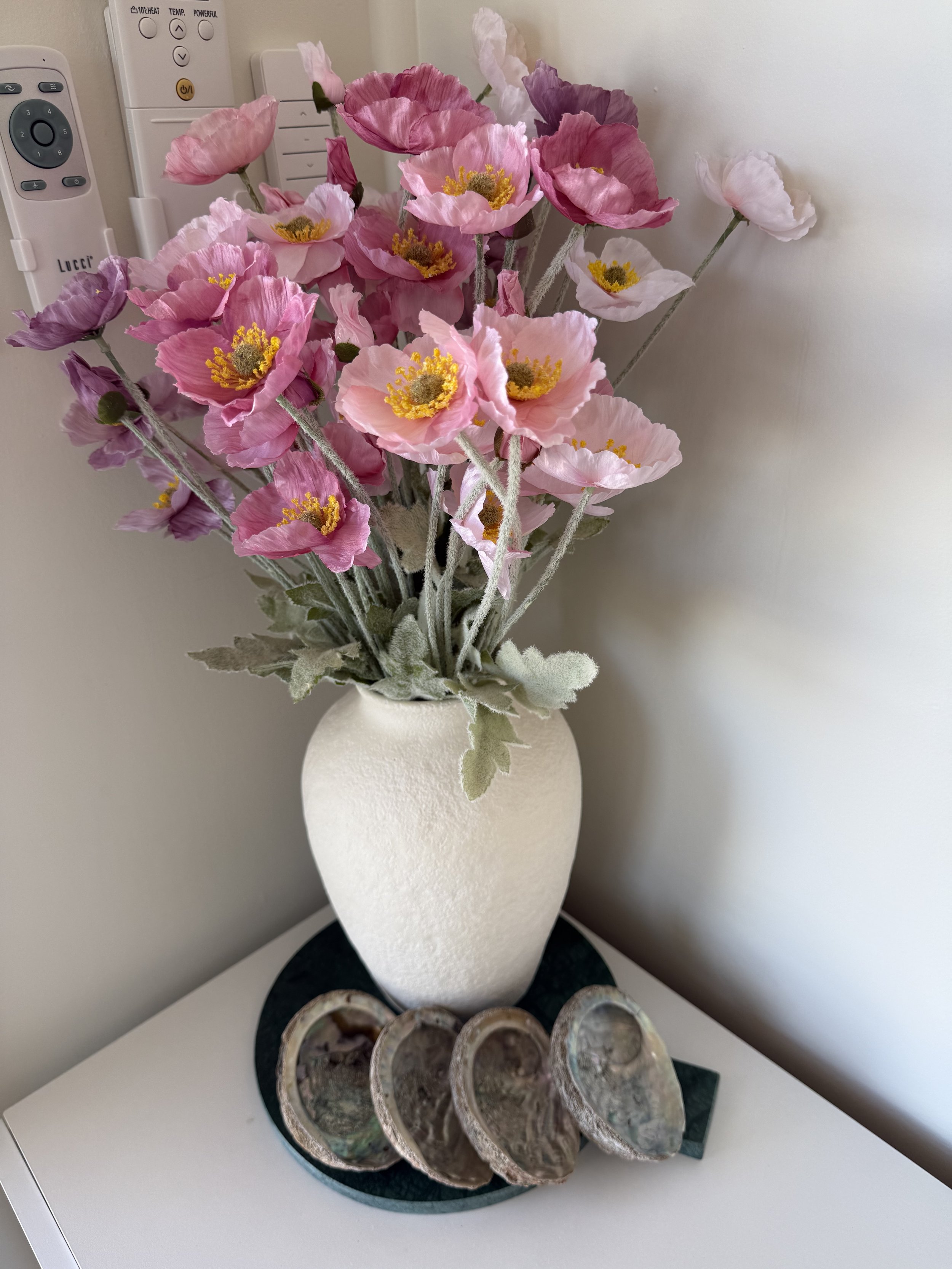 A white textured ceramic vase with artificial pink and purple poppy flowers with yellow centers, placed on a round black base on a white surface, with three seashells arranged in front of it.