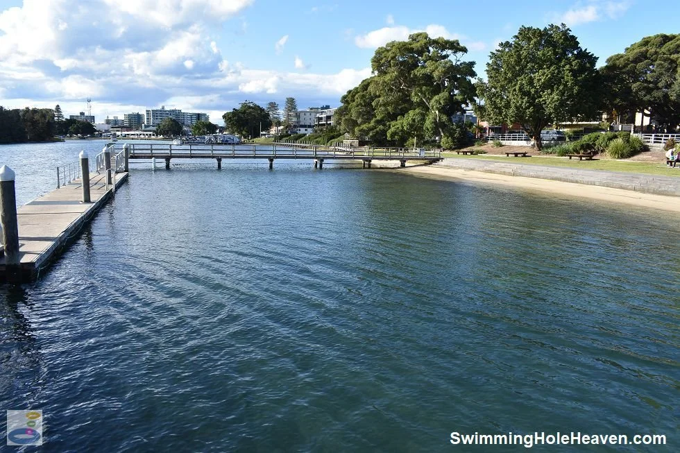 A waterfront park with a dock extending into the water, trees lining the shore, and buildings in the background under a partly cloudy sky.