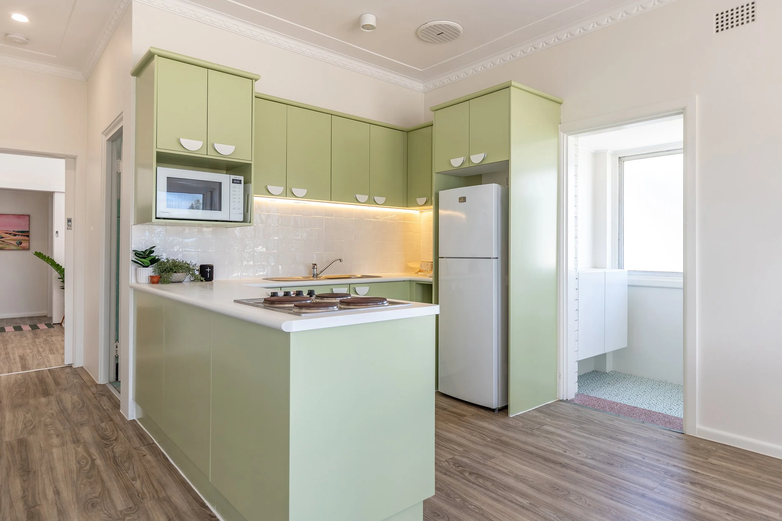 A kitchen with light green cabinets, white countertops, a white refrigerator, a microwave, potted plants, a white tiled backsplash, and a small laundry or utility area with a window.
