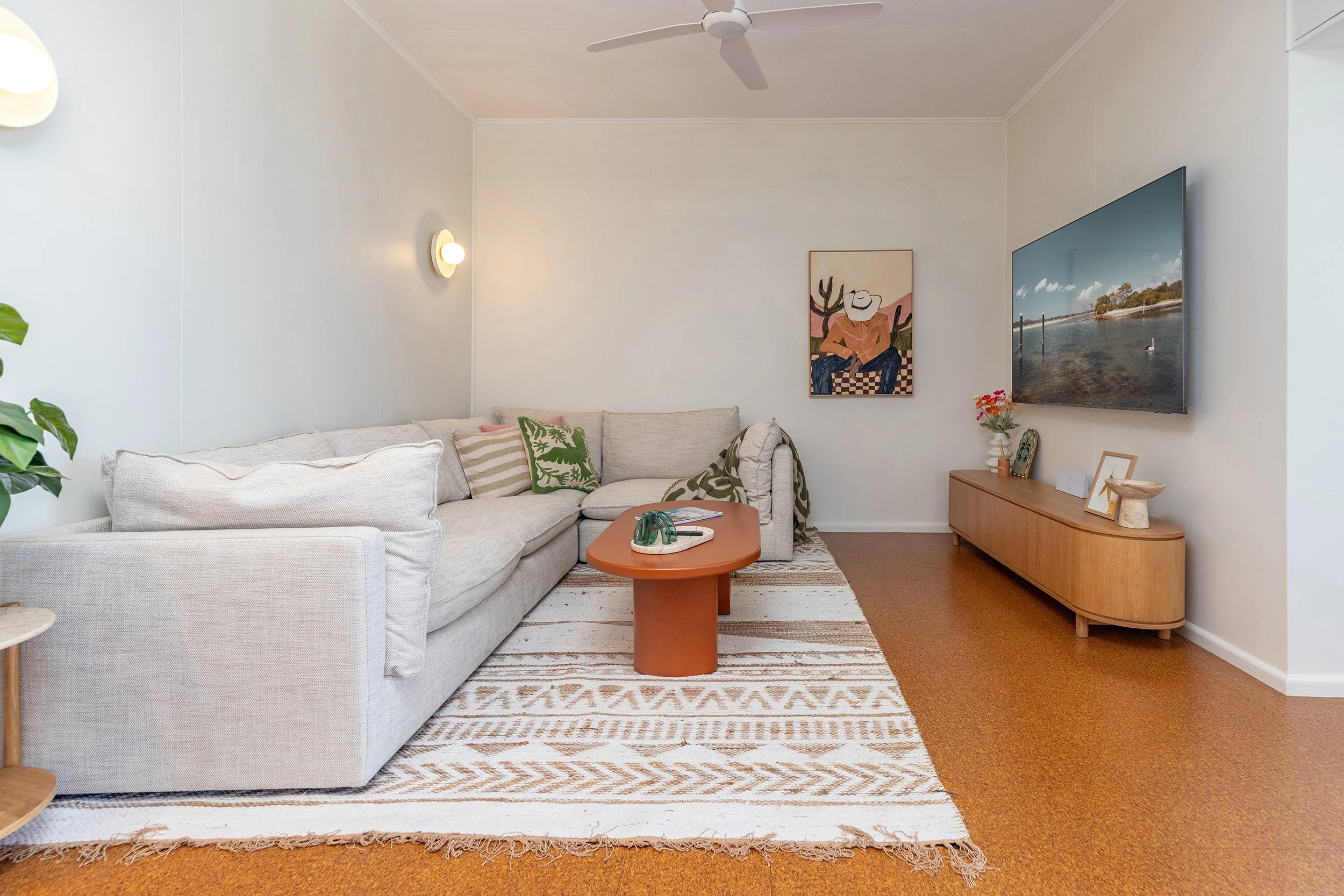 Living room with a large beige sofa, a rust-colored oval coffee table, a patterned rug, a wooden TV stand, a flat-screen TV, wall art, a ceiling fan, and decor objects.