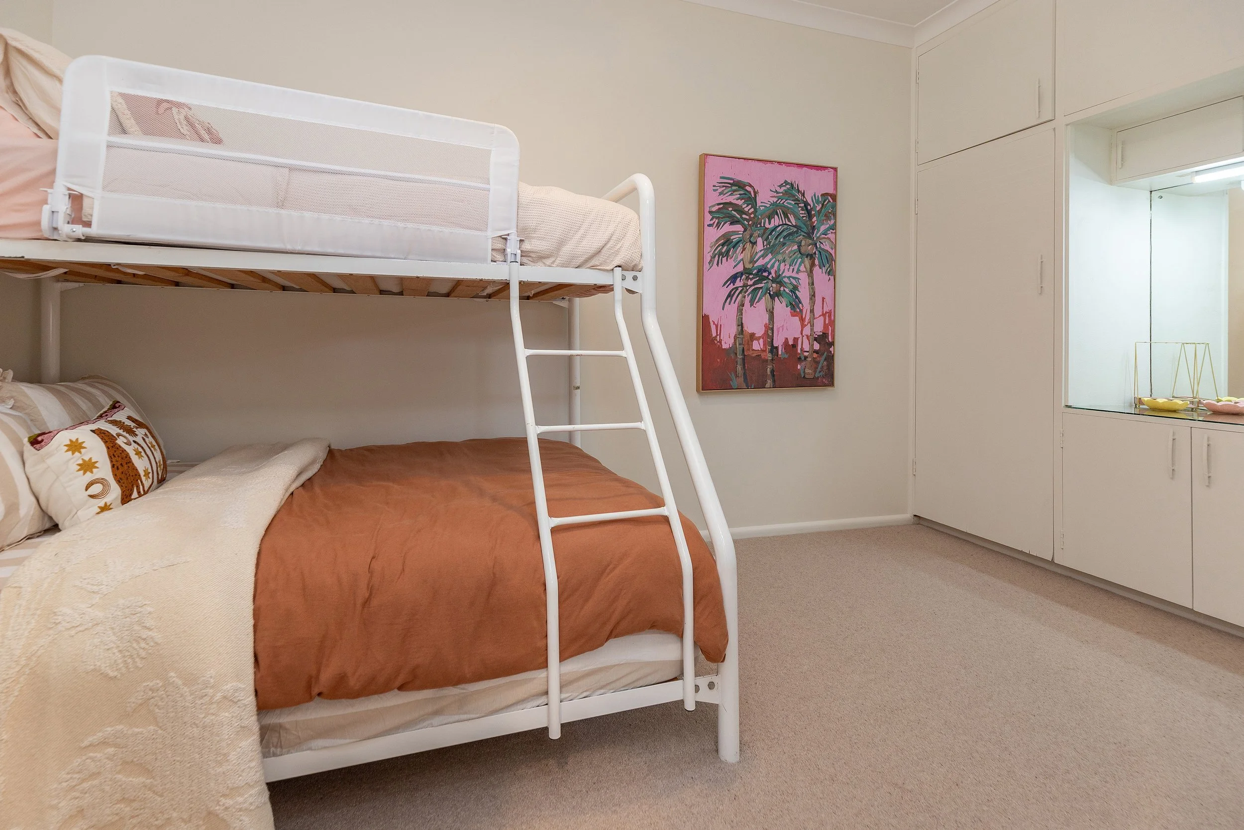 Bedroom with a white bunk bed, brown bedding, decorative pillows, a colorful painting of palm trees on the wall, and a window with built-in cabinets.