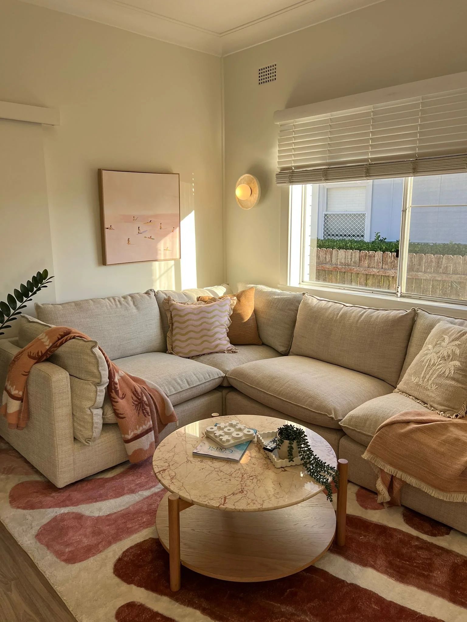 Living room with beige sectional sofa, pink and brown throw pillows, a pink patterned blanket, a round marble-top coffee table, and a colorful rug. Sunlight streams through a window with white blinds, illuminating the room.