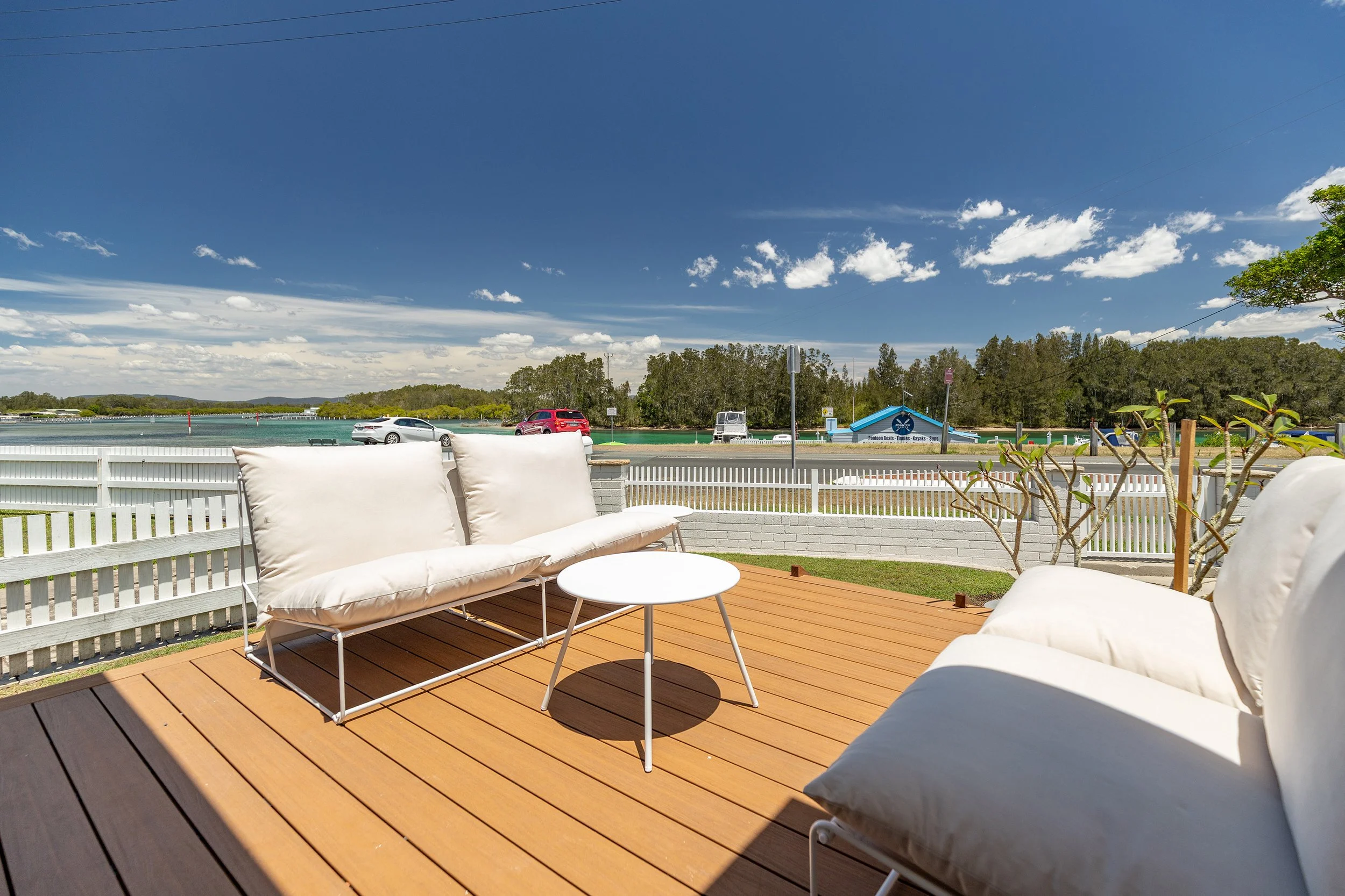 A view of an outdoor wooden deck with white cushioned patio furniture, overlooking a lake or river with a blue sky and some clouds, and a white fence in the background.