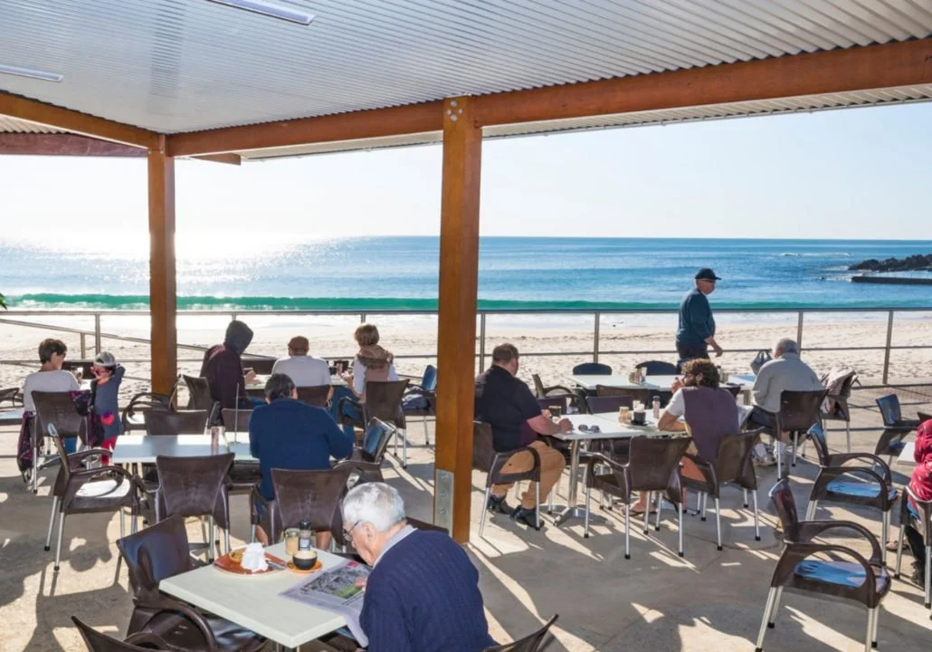People dining at an outdoor restaurant overlooking the beach with blue ocean and sunny sky.